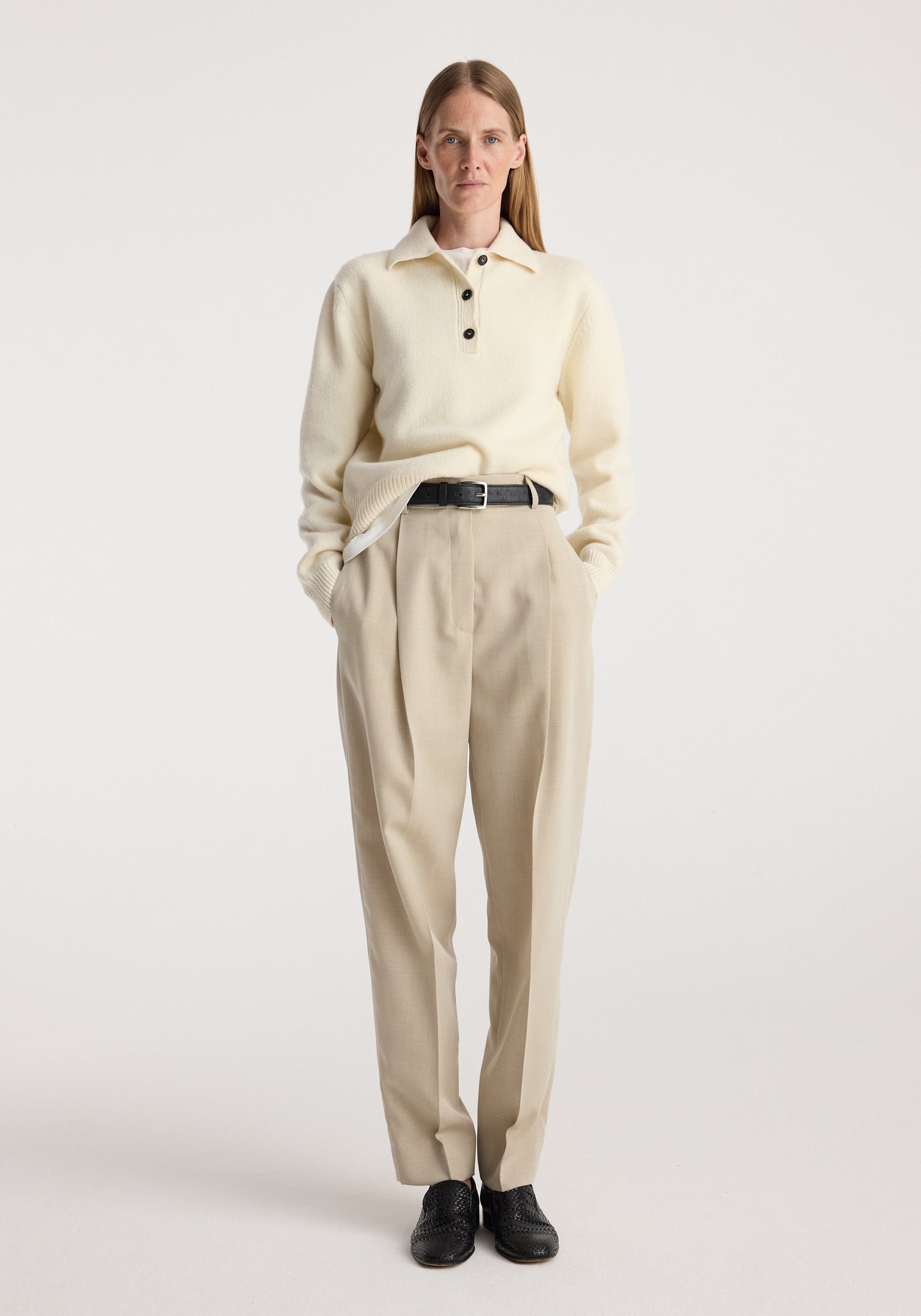 Woman wearing Daria - Single pleat straight leg trousers | butter in a beige tone, paired with an off-white long-sleeve sweater with buttons and black shoes.