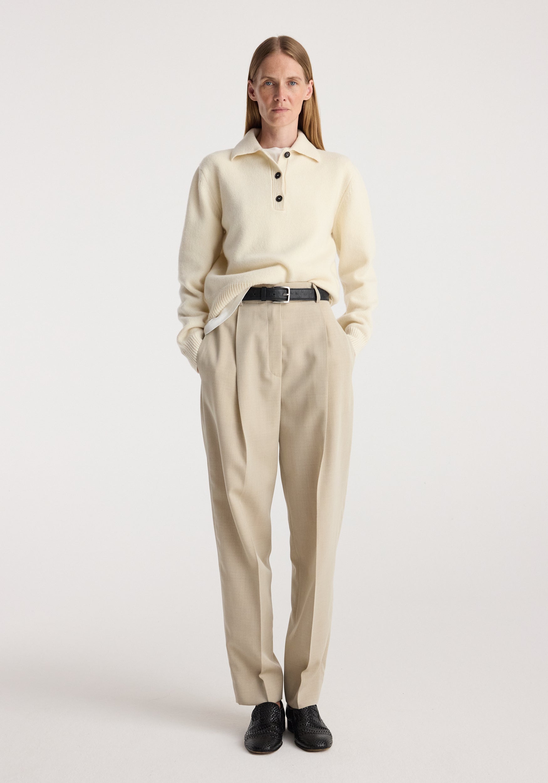 Woman wearing Daria - Single pleat straight leg trousers | butter in a beige tone, paired with an off-white long-sleeve sweater with buttons and black shoes.