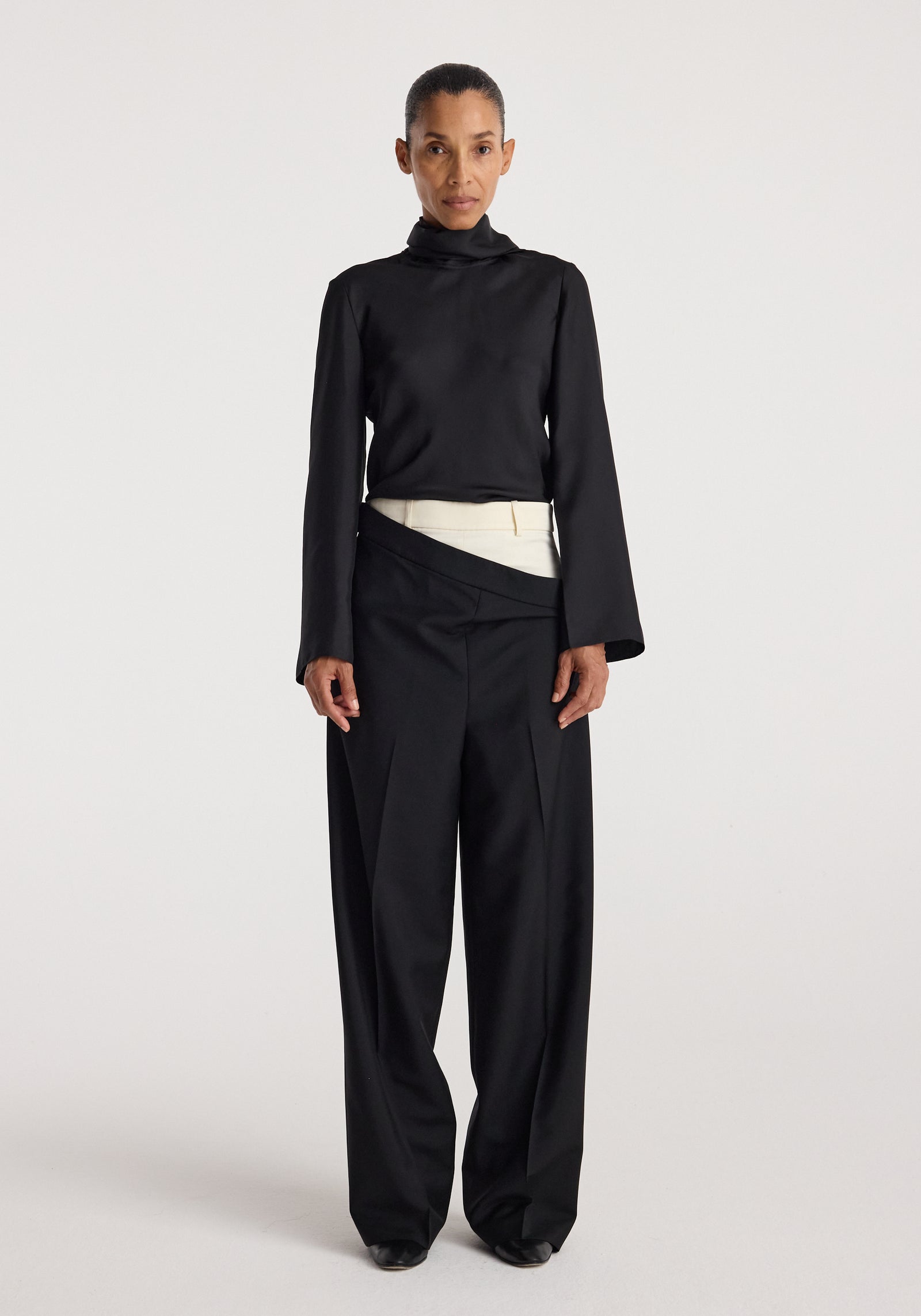 Front view of a woman wearing a black high-neck long sleeve top paired with Double waistband trousers | black featuring a unique double waistband design with an overlapping cream-colored layer.
