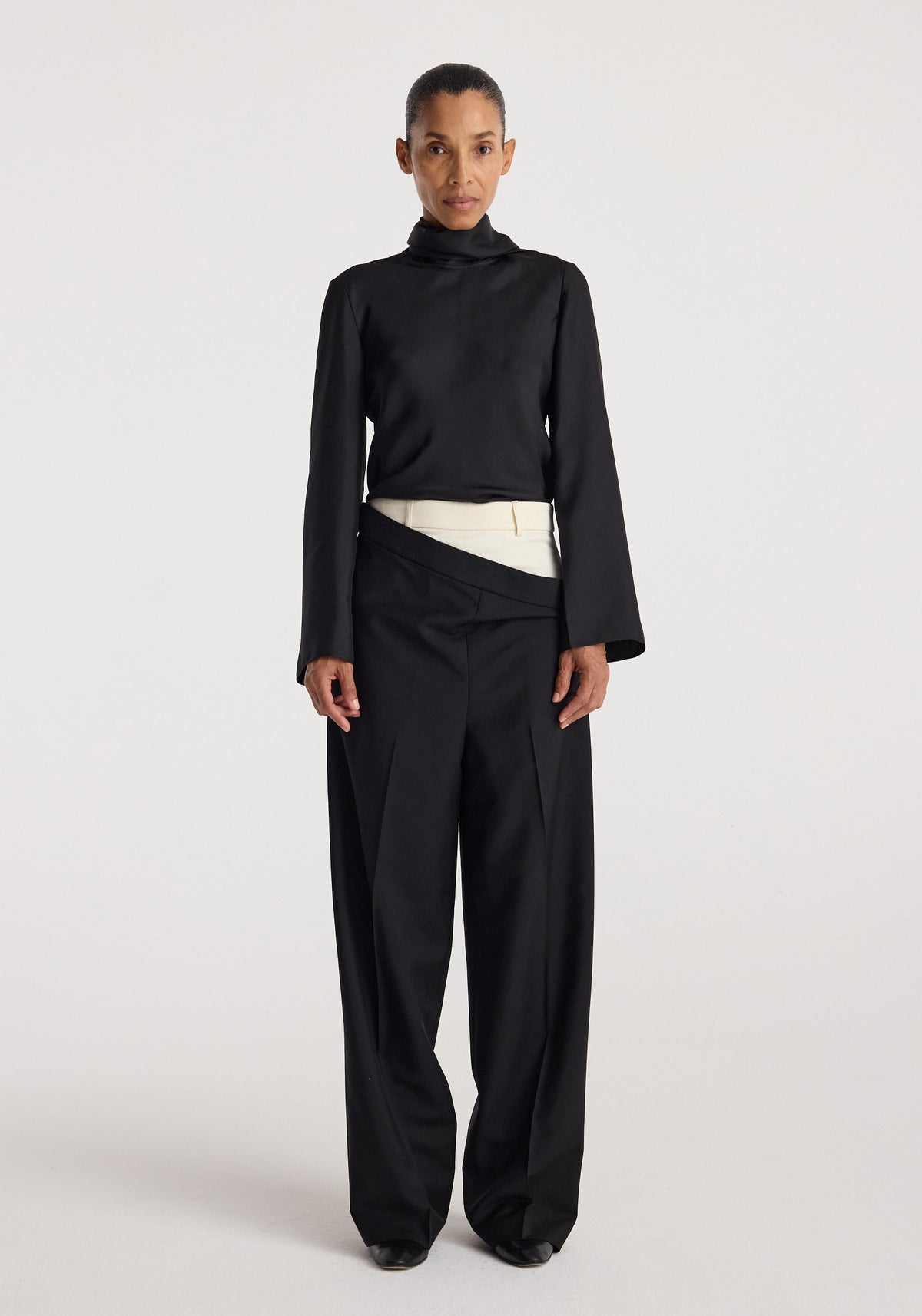 Front view of a woman wearing a black high-neck long sleeve top paired with Double waistband trousers | black featuring a unique double waistband design with an overlapping cream-colored layer.