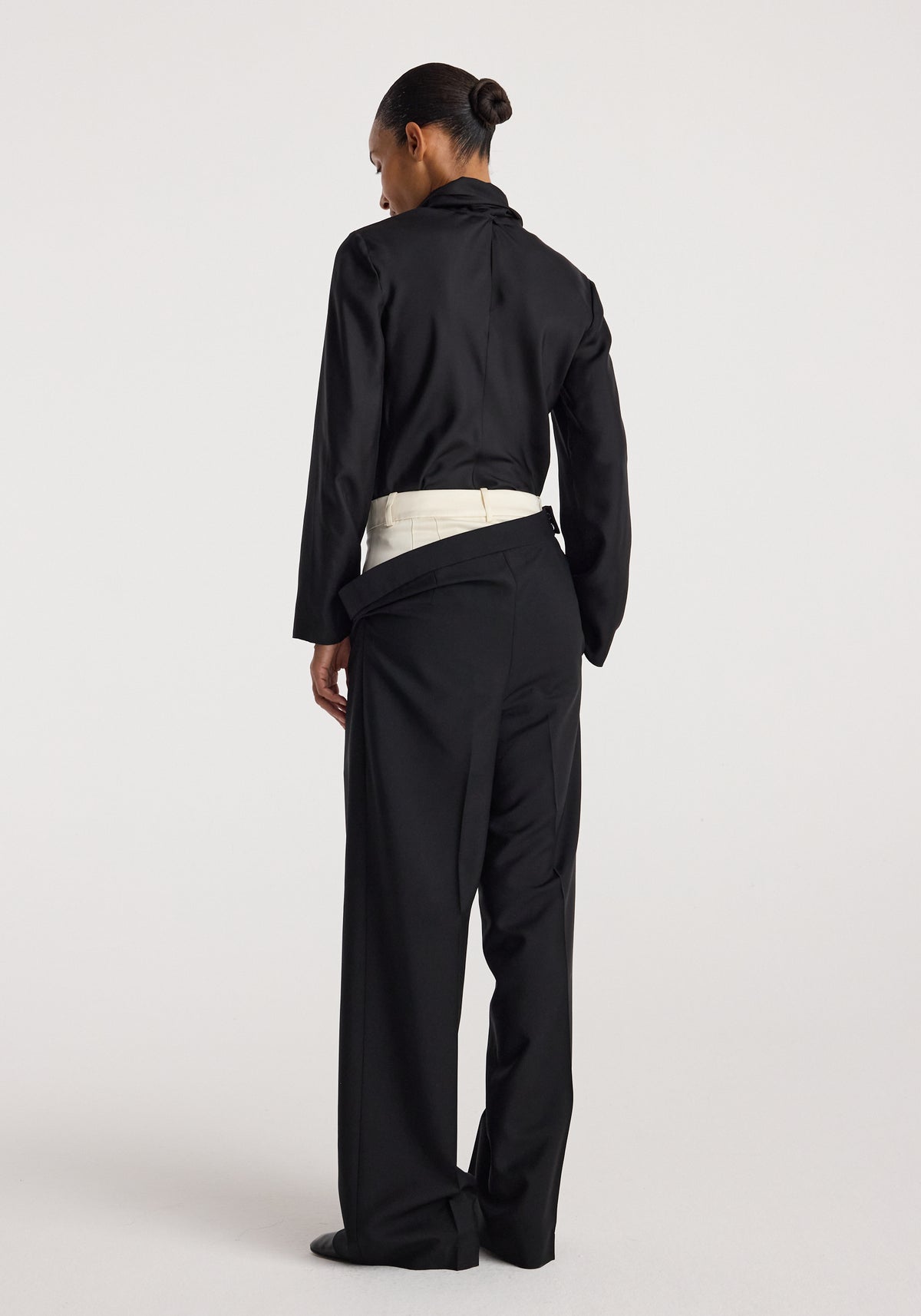 Rear view of a woman wearing black Double waistband trousers | black with an inner cream waistband visible, paired with a black long-sleeve top and hair in a bun.