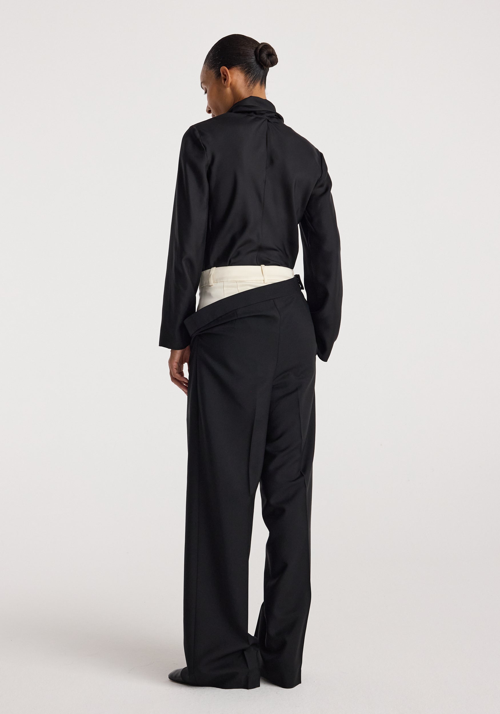 Rear view of a woman wearing black Double waistband trousers | black with an inner cream waistband visible, paired with a black long-sleeve top and hair in a bun.