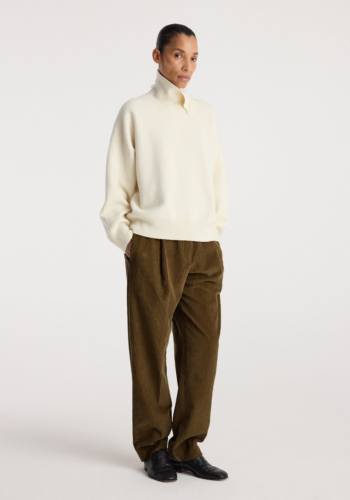 A model wearing Daria - Single pleat straight leg corduroy trousers | caramel paired with a cream high-neck sweater and black shoes, standing against a plain background.