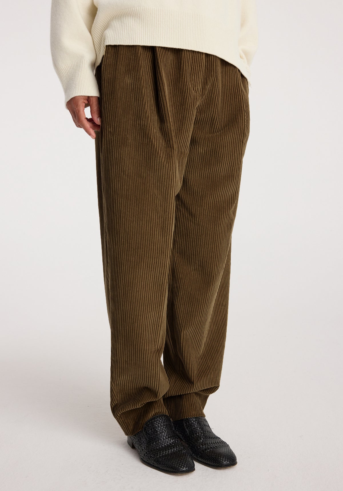 Close-up of a person wearing Daria - Single pleat straight leg corduroy trousers | caramel paired with a cream sweater and black woven shoes against a plain background.