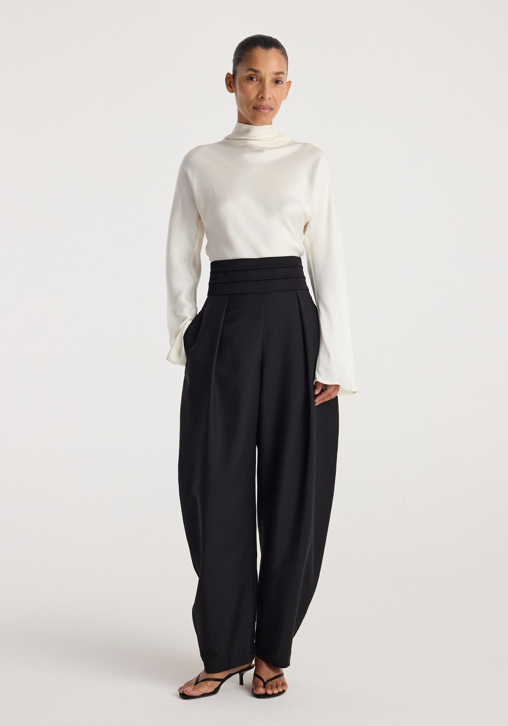 Woman wearing a white high-neck blouse paired with black Puck - Sculptural cumberband trousers | black featuring a high, layered waistband and wide legs, styled with black heeled sandals.