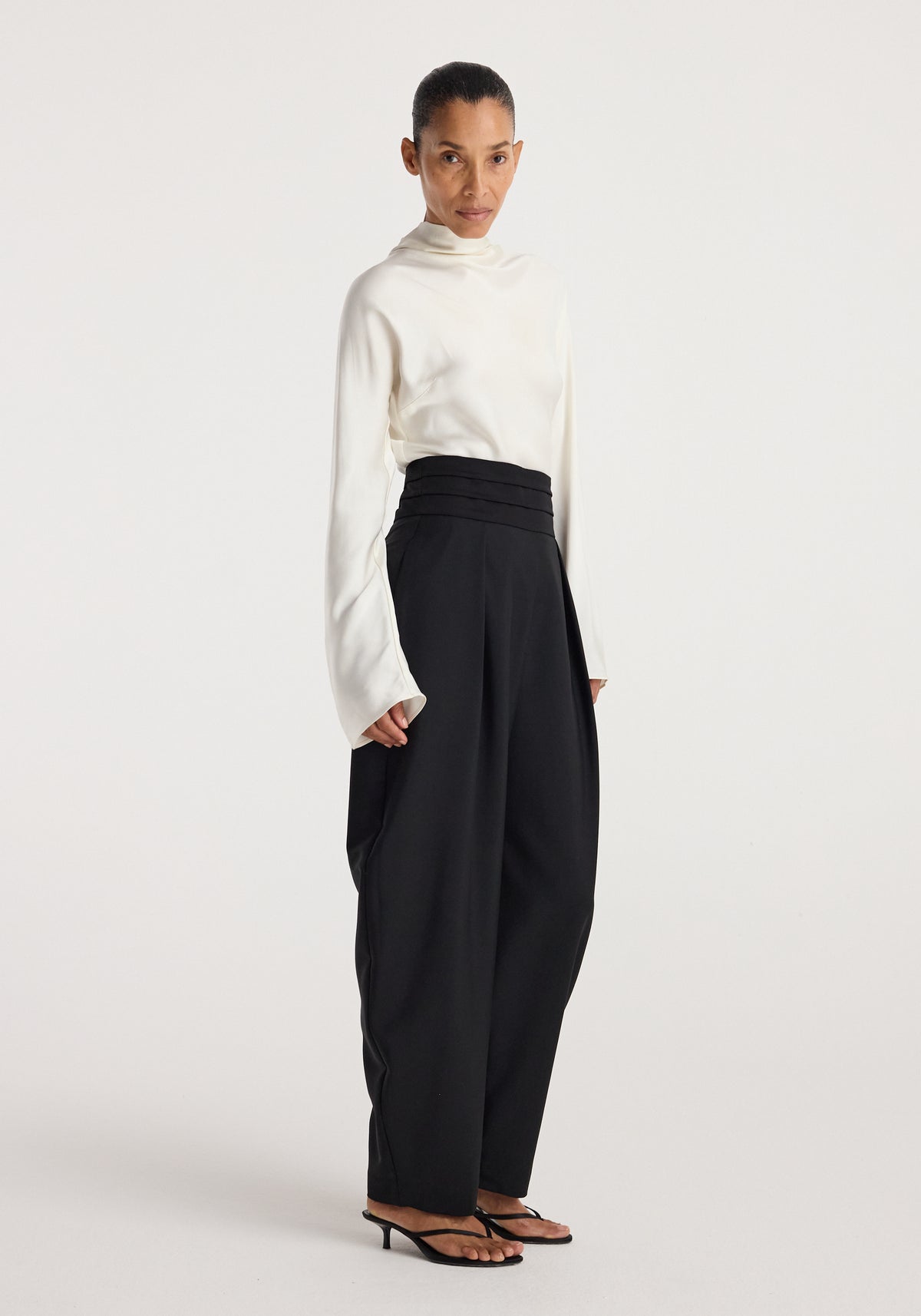 Model wearing black Puck - Sculptural cumberband trousers | black paired with a white long-sleeve top and black open-toe heels against a plain background.
