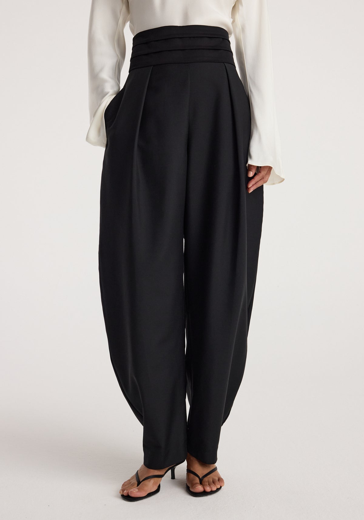 Front view of a model wearing Puck - Sculptural cumberband trousers | black with a high waistband, pleated detailing, and a relaxed, tapered fit paired with a white long-sleeve blouse and black heeled sandals.