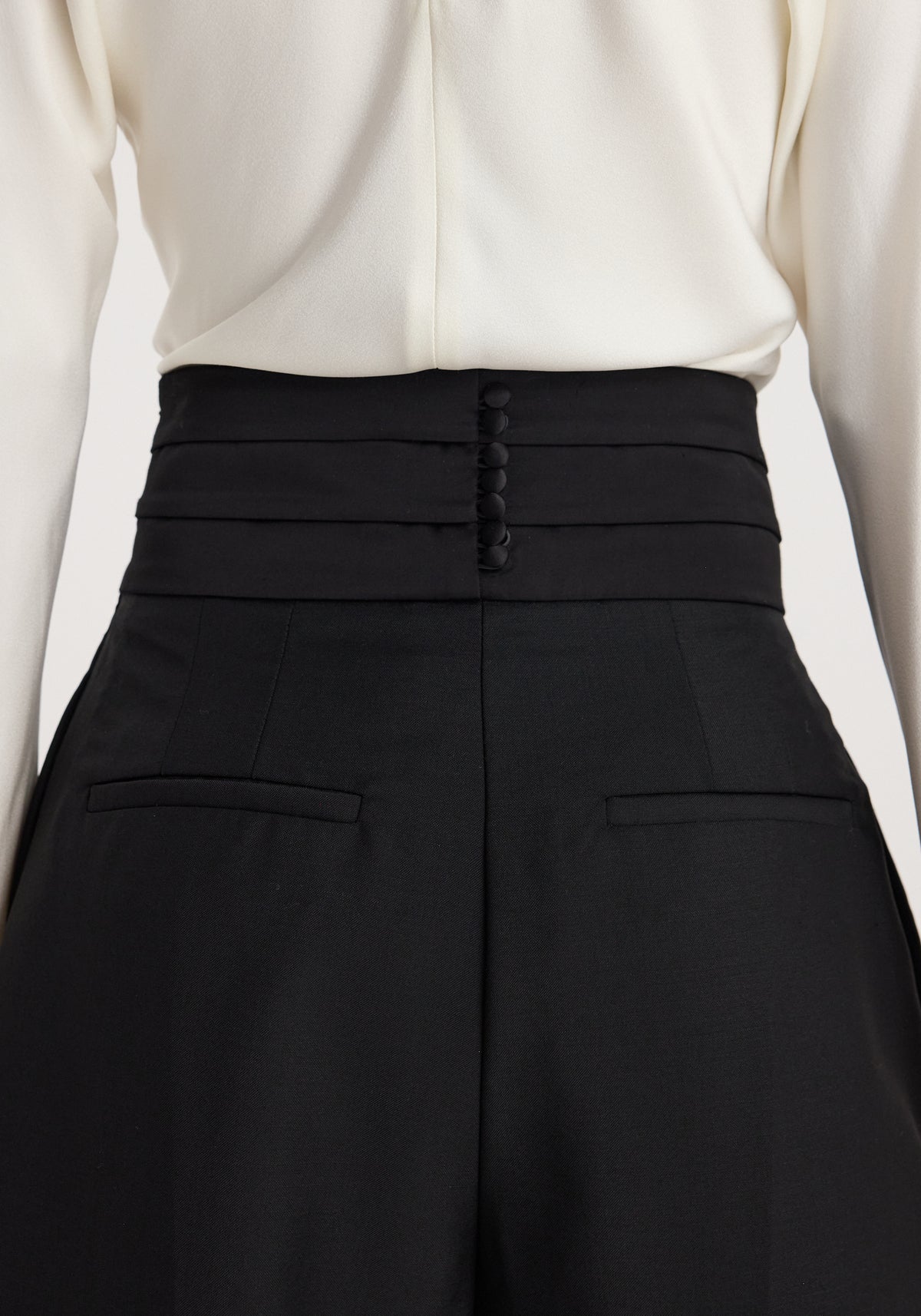 Back view of a person wearing black Puck - Sculptural cumberband trousers | black with button detail and a high waist paired with a white top.