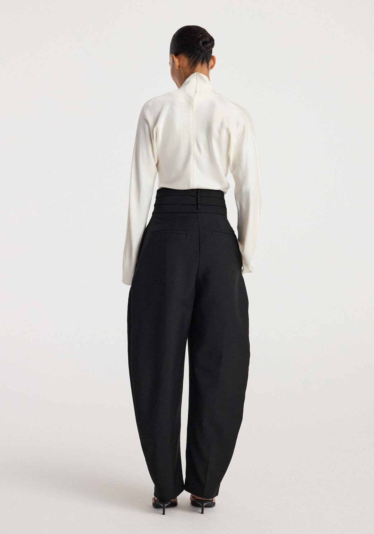 Rear view of a woman wearing black Puck - Sculptural cumberband trousers | black paired with a white long-sleeve top and black heeled shoes against a plain light background.