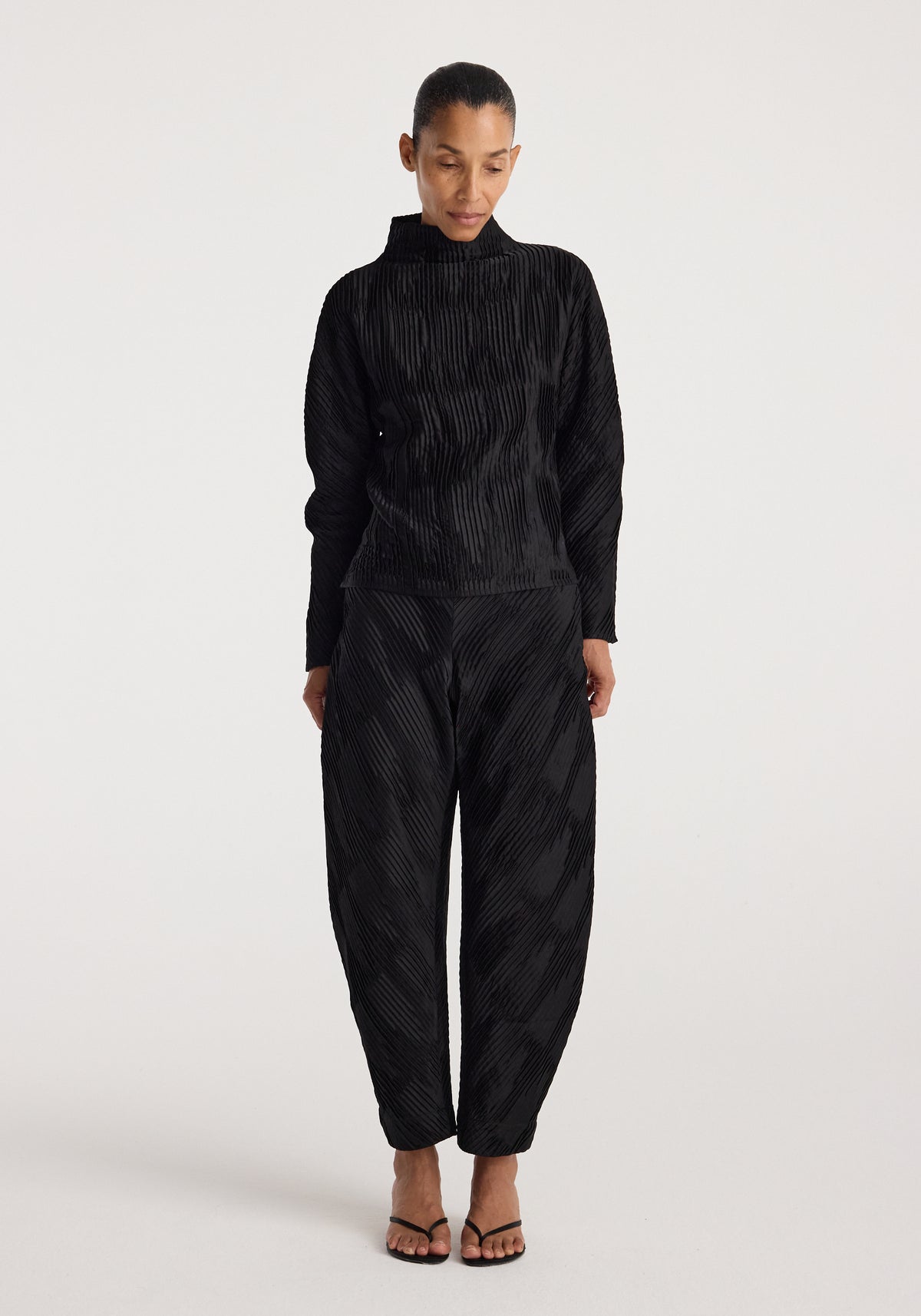 Woman wearing black plissé satin trousers | black with matching long-sleeve top and black open-toe heels against a plain white background.