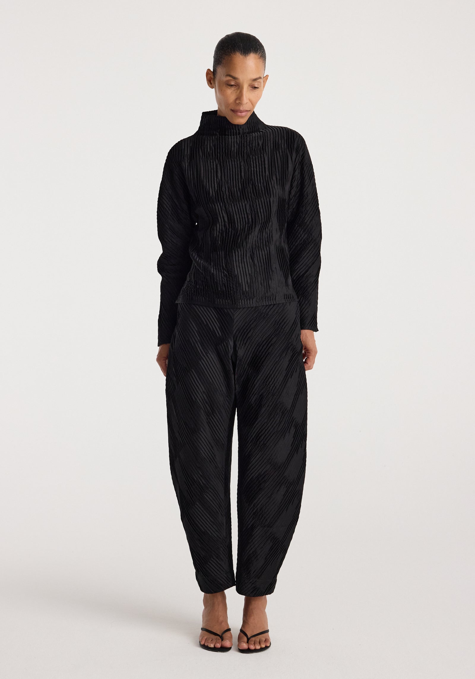 Woman wearing black plissé satin trousers | black with matching long-sleeve top and black open-toe heels against a plain white background.
