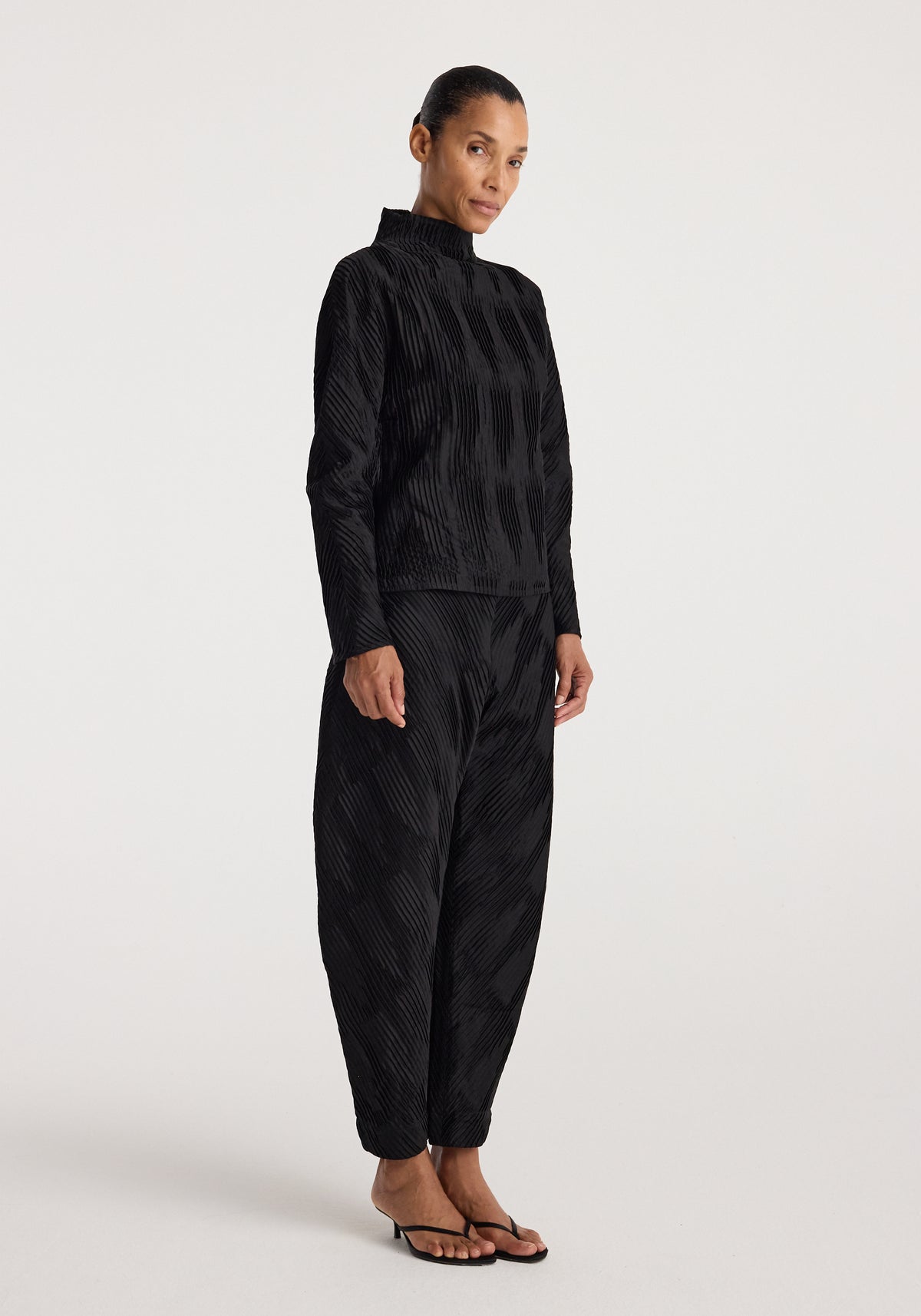 Woman wearing black plissé satin trousers | black with matching long-sleeve top, standing against a plain light background, styled with black sandals.
