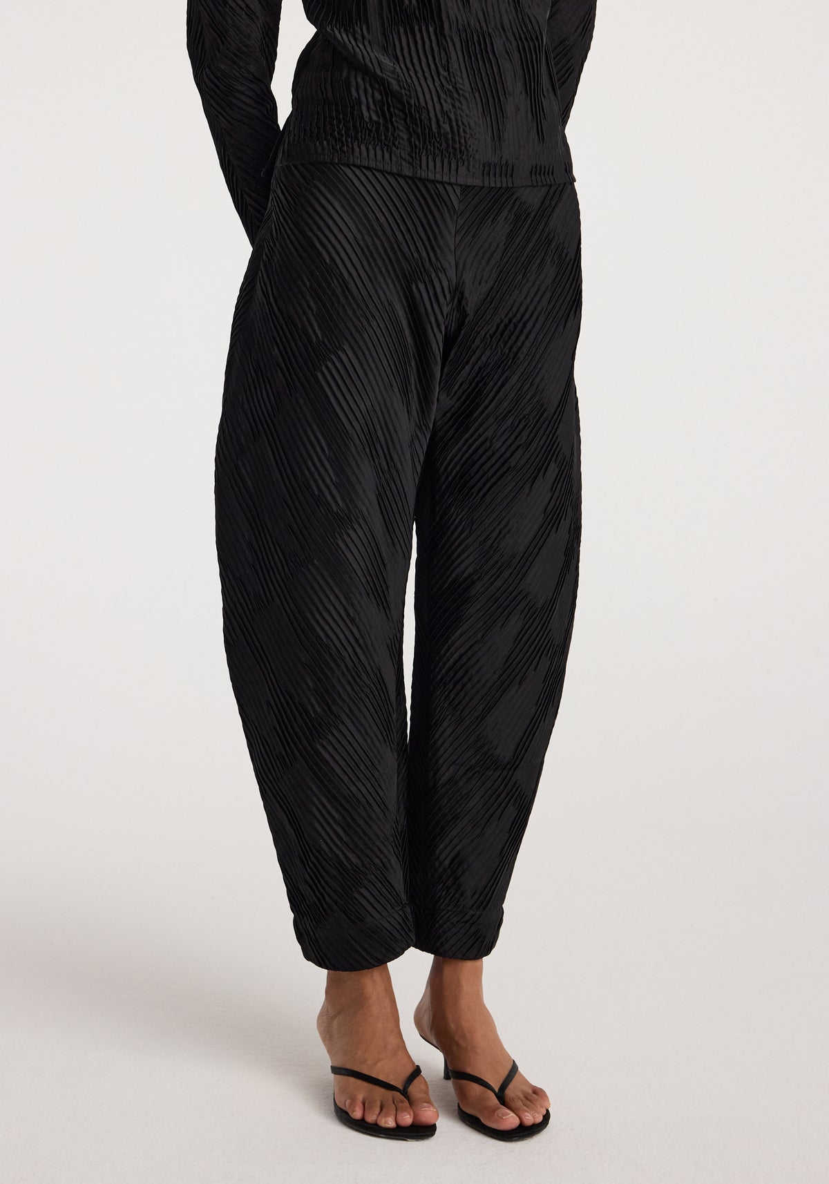 Close-up of a person wearing black Plissé satin trousers with a textured, pleated design paired with black open-toe heeled sandals against a plain background.