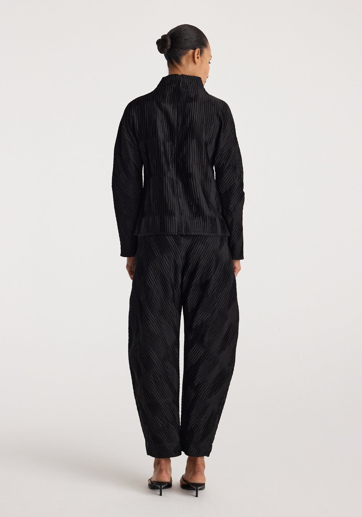 Rear view of a person wearing black Plissé satin trousers | black with a matching long-sleeve top, standing against a plain white background.