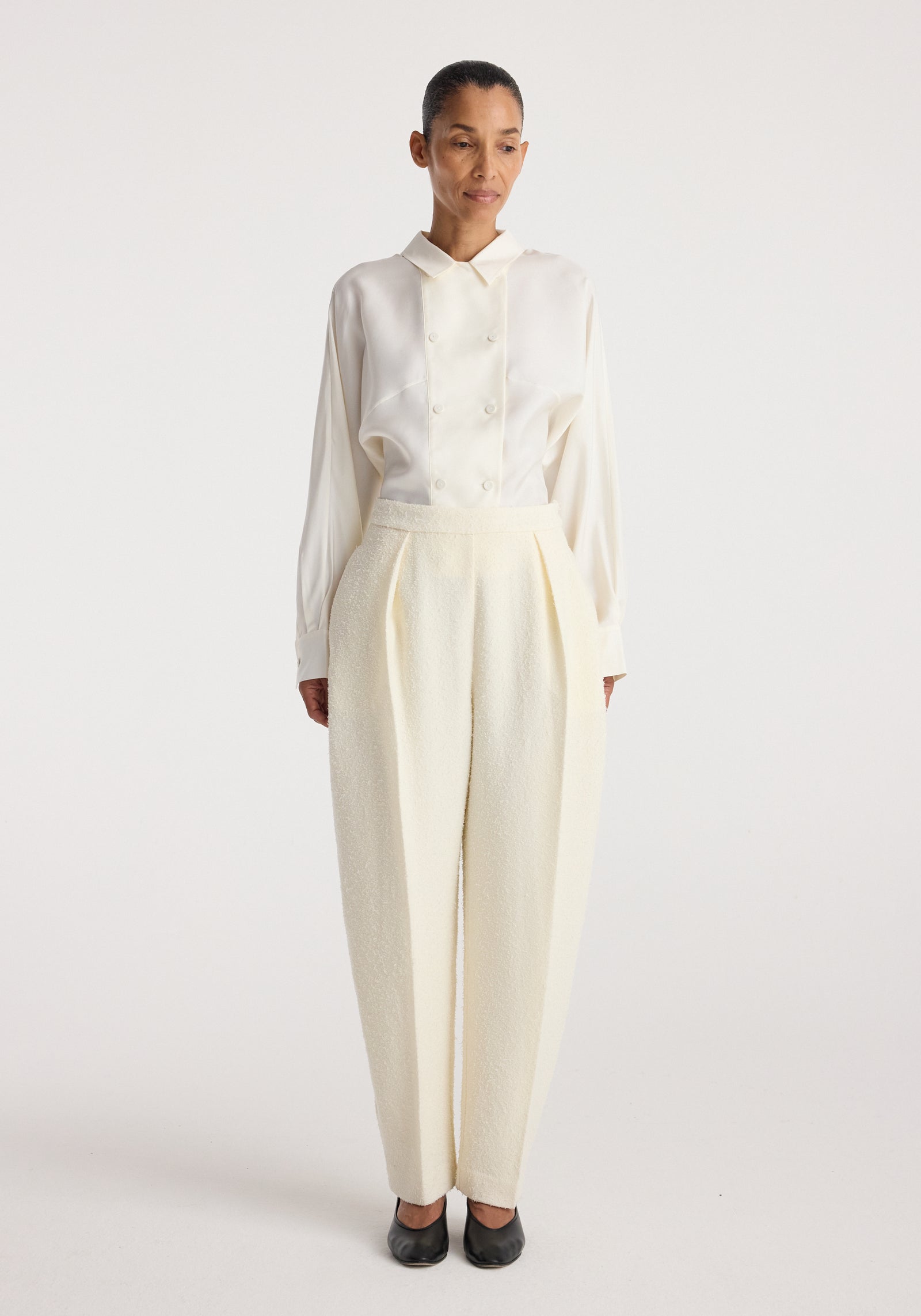 Woman wearing Puck - Sculptural bouclé tailored trousers | cream with a tucked-in long-sleeve cream blouse and black shoes, standing against a plain light background.