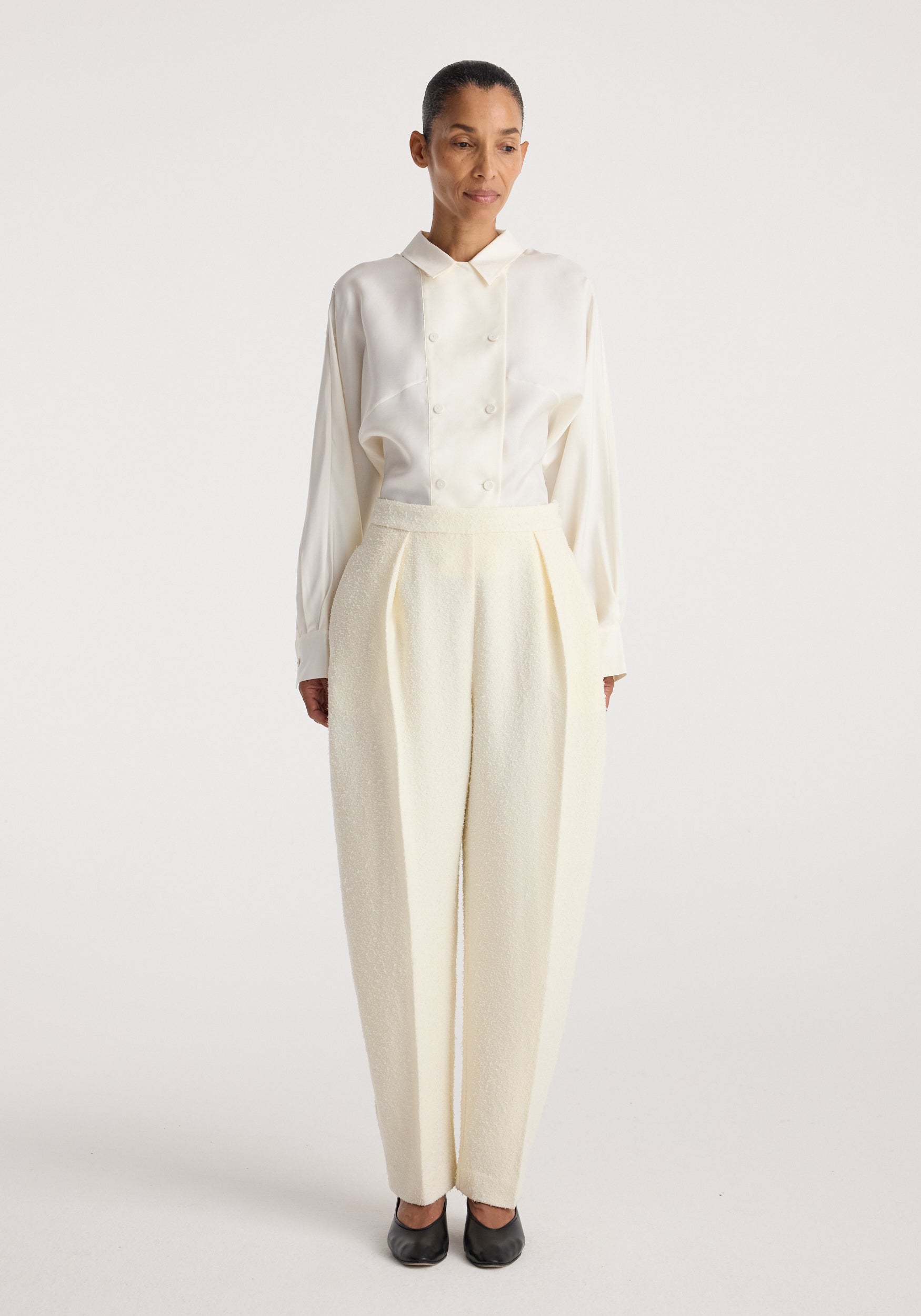 Woman wearing Puck - Sculptural bouclé tailored trousers | cream with a tucked-in long-sleeve cream blouse and black shoes, standing against a plain light background.