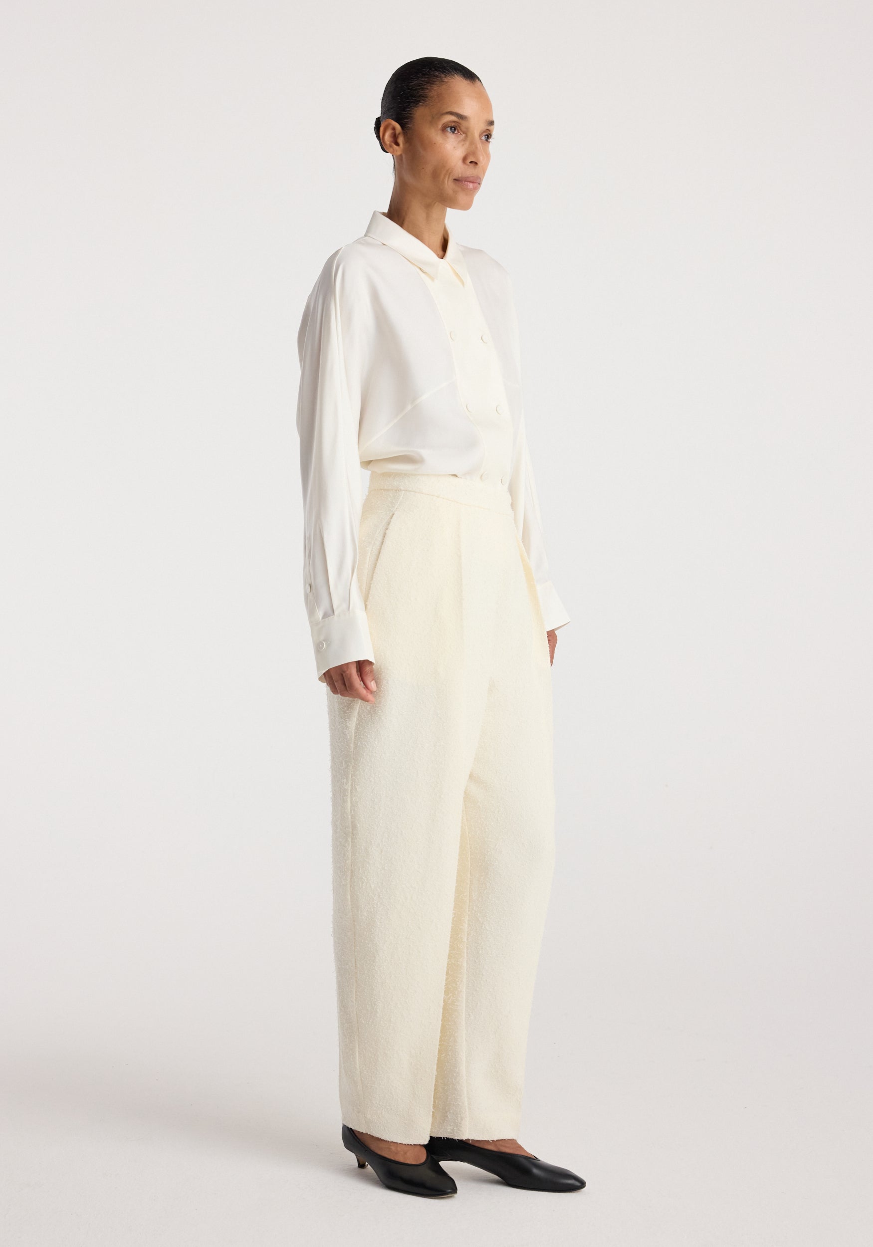 Woman wearing Puck - Sculptural bouclé tailored trousers | cream with a white button-up shirt and black low-heeled shoes, standing against a plain background.
