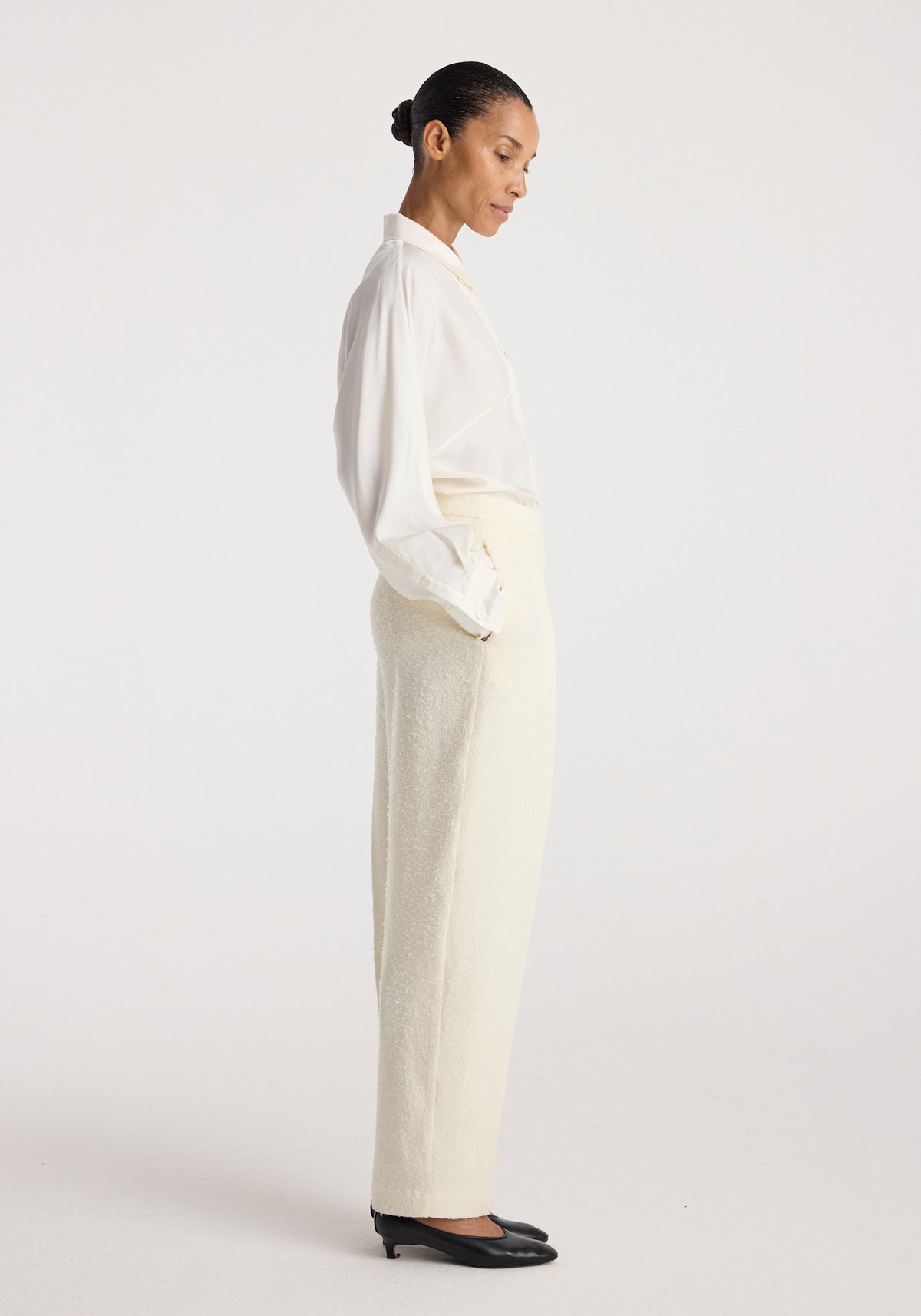 Model wearing Puck - Sculptural bouclé tailored trousers | cream paired with a white blouse and black heels, shown in a side profile against a plain background.