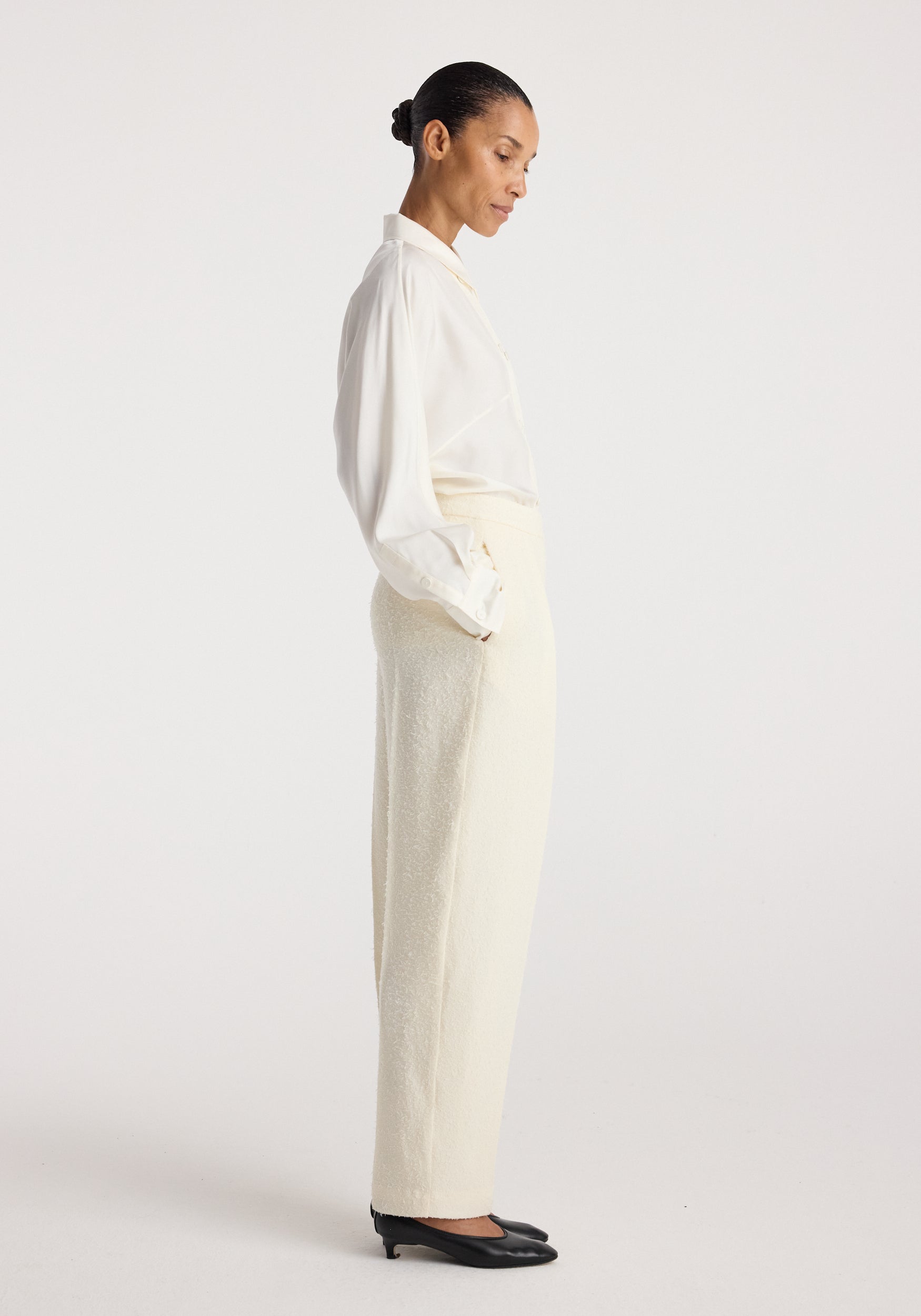 Model wearing Puck - Sculptural bouclé tailored trousers | cream paired with a white blouse and black heels, shown in a side profile against a plain background.