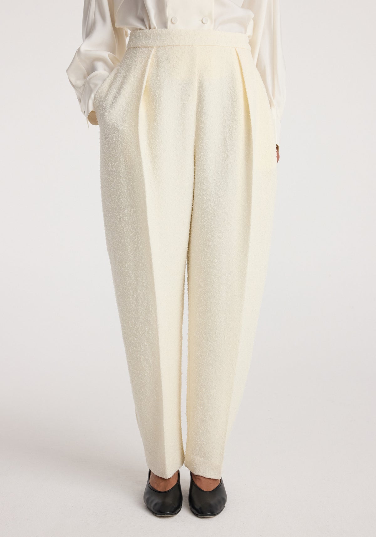 Front view of cream-colored Puck - Sculptural bouclé tailored trousers | cream worn with hands in pockets, paired with black shoes and a white long-sleeve top.