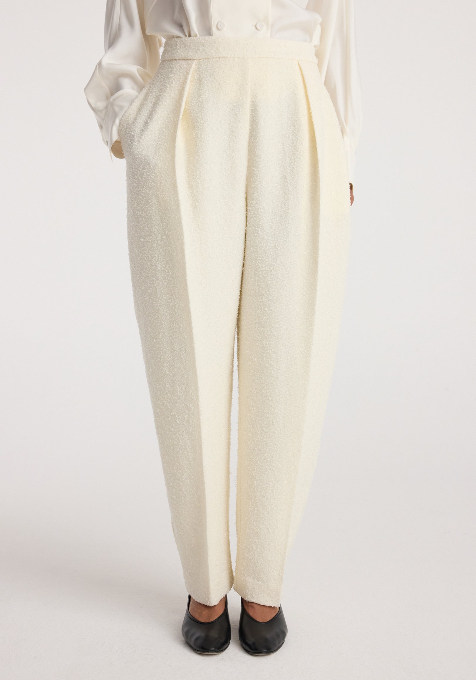 Front view of cream-colored Puck - Sculptural bouclé tailored trousers | cream worn with hands in pockets, paired with black shoes and a white long-sleeve top.