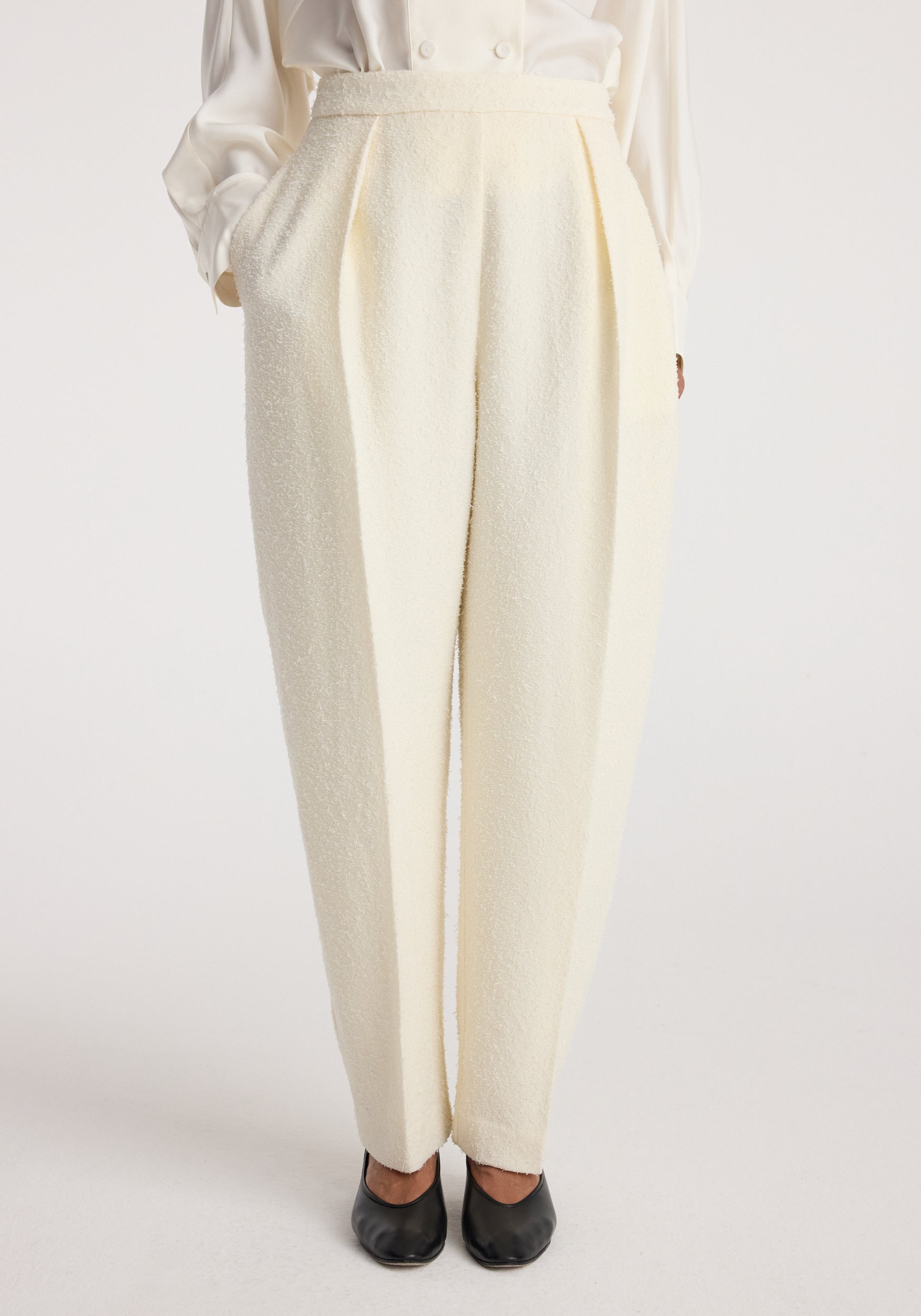Front view of cream-colored Puck - Sculptural bouclé tailored trousers | cream worn with hands in pockets, paired with black shoes and a white long-sleeve top.