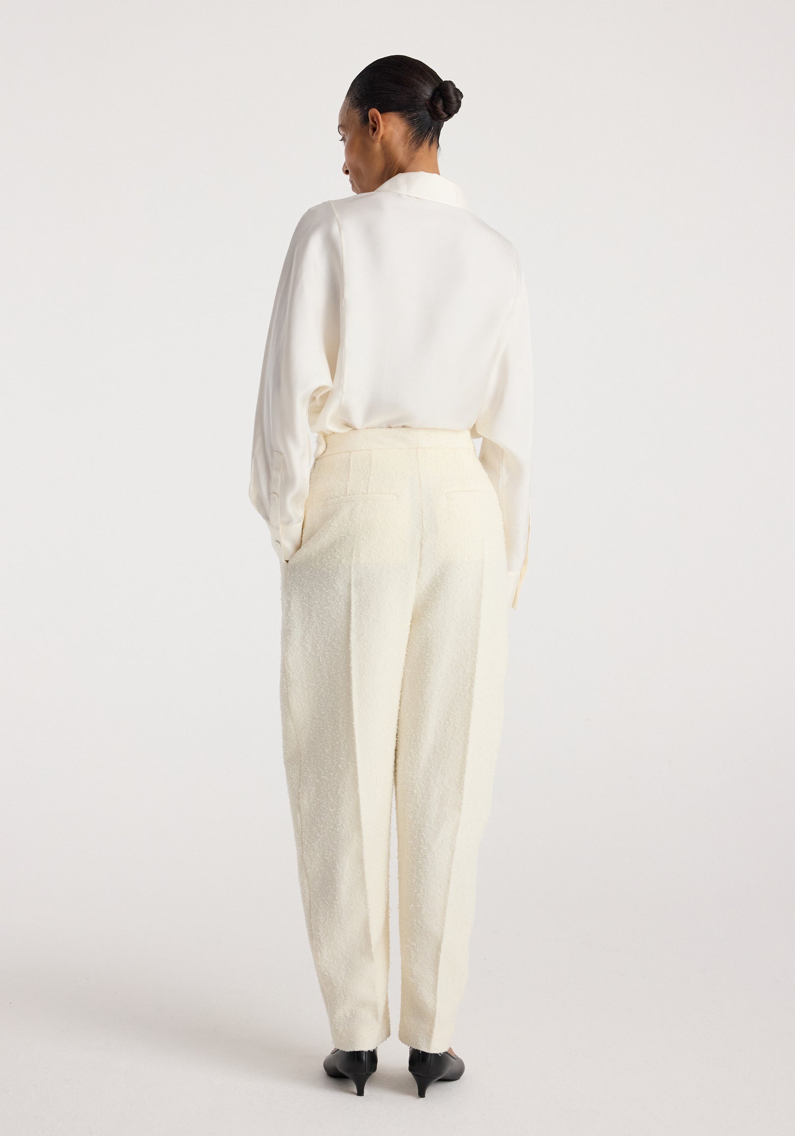 Rear view of a woman wearing Puck - Sculptural bouclé tailored trousers | cream, paired with a white blouse and black heels, standing against a plain background.