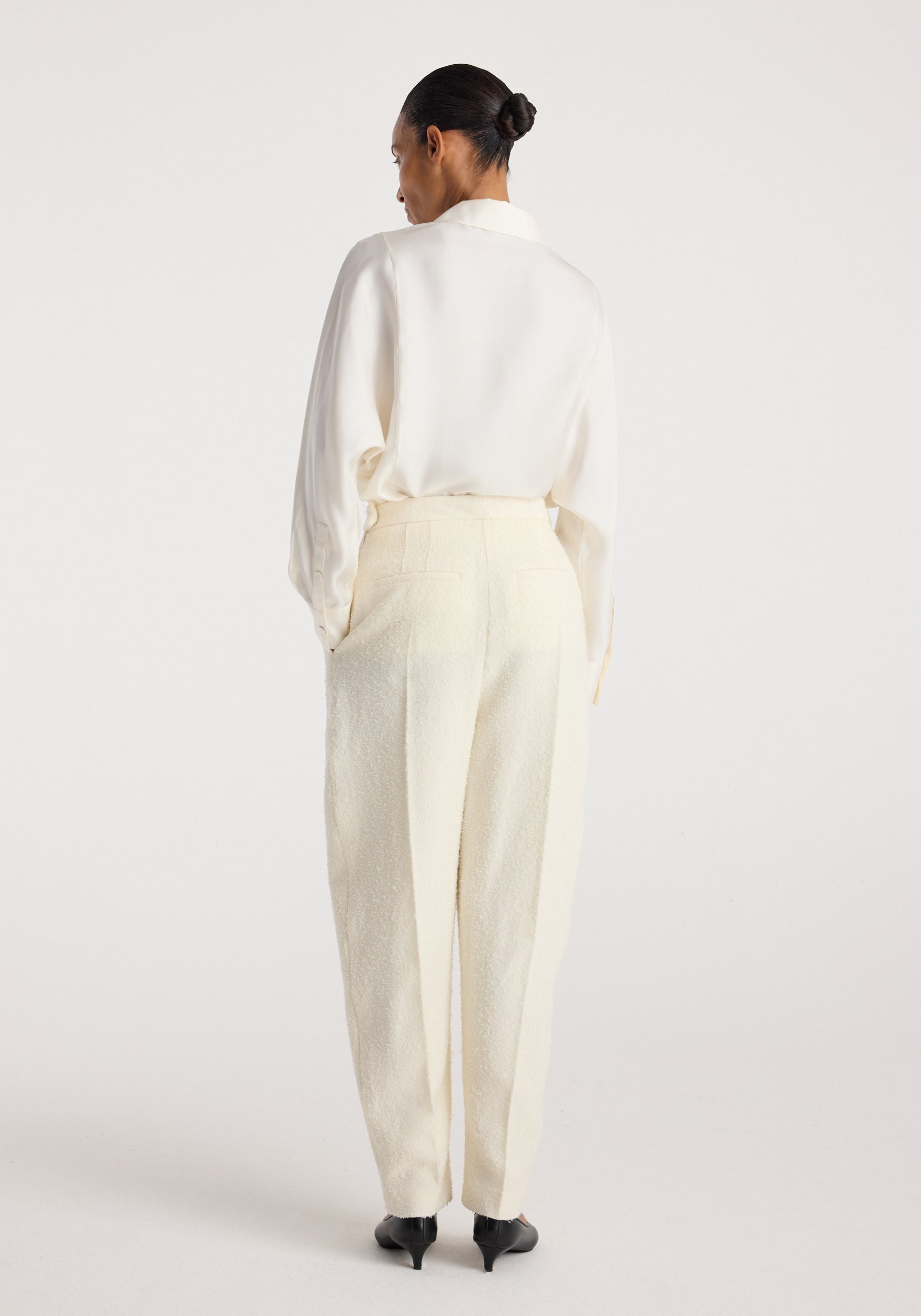Rear view of a woman wearing Puck - Sculptural bouclé tailored trousers | cream, paired with a white blouse and black heels, standing against a plain background.