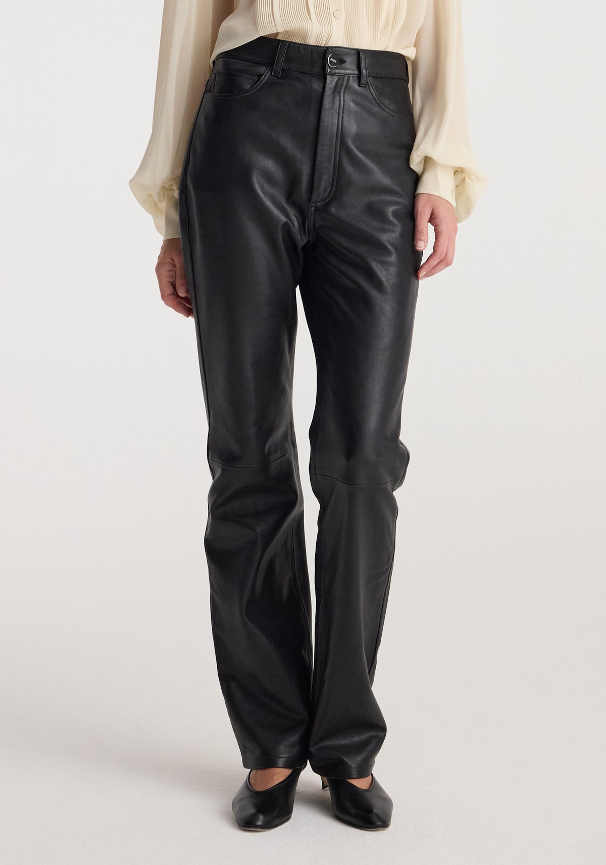 Woman wearing high-waist straight leather trousers | black paired with a light, long-sleeve blouse and black shoes, standing against a plain background.