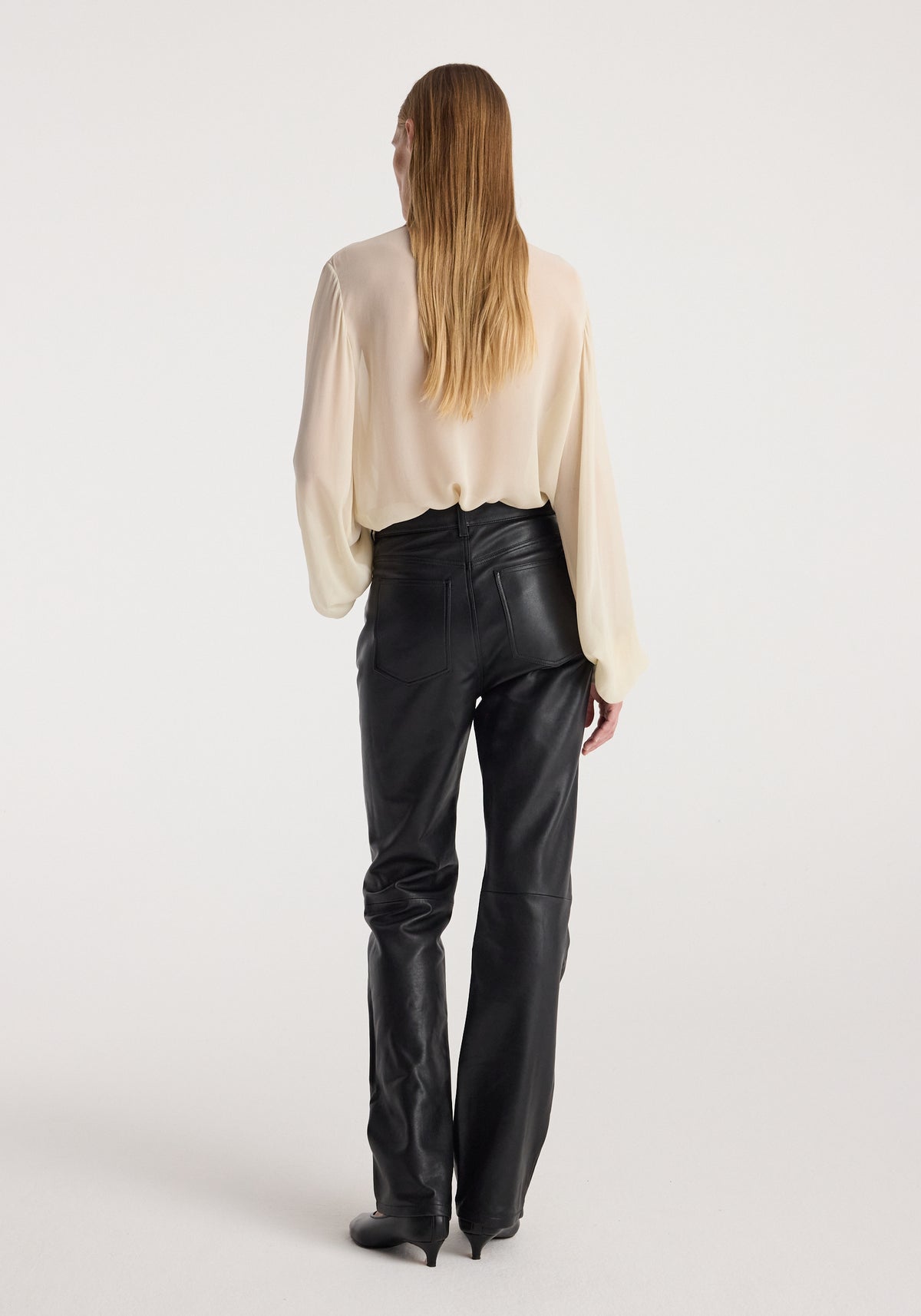 Woman seen from behind wearing a light blouse paired with high-waist straight leather trousers | black and black heeled shoes against a plain background.