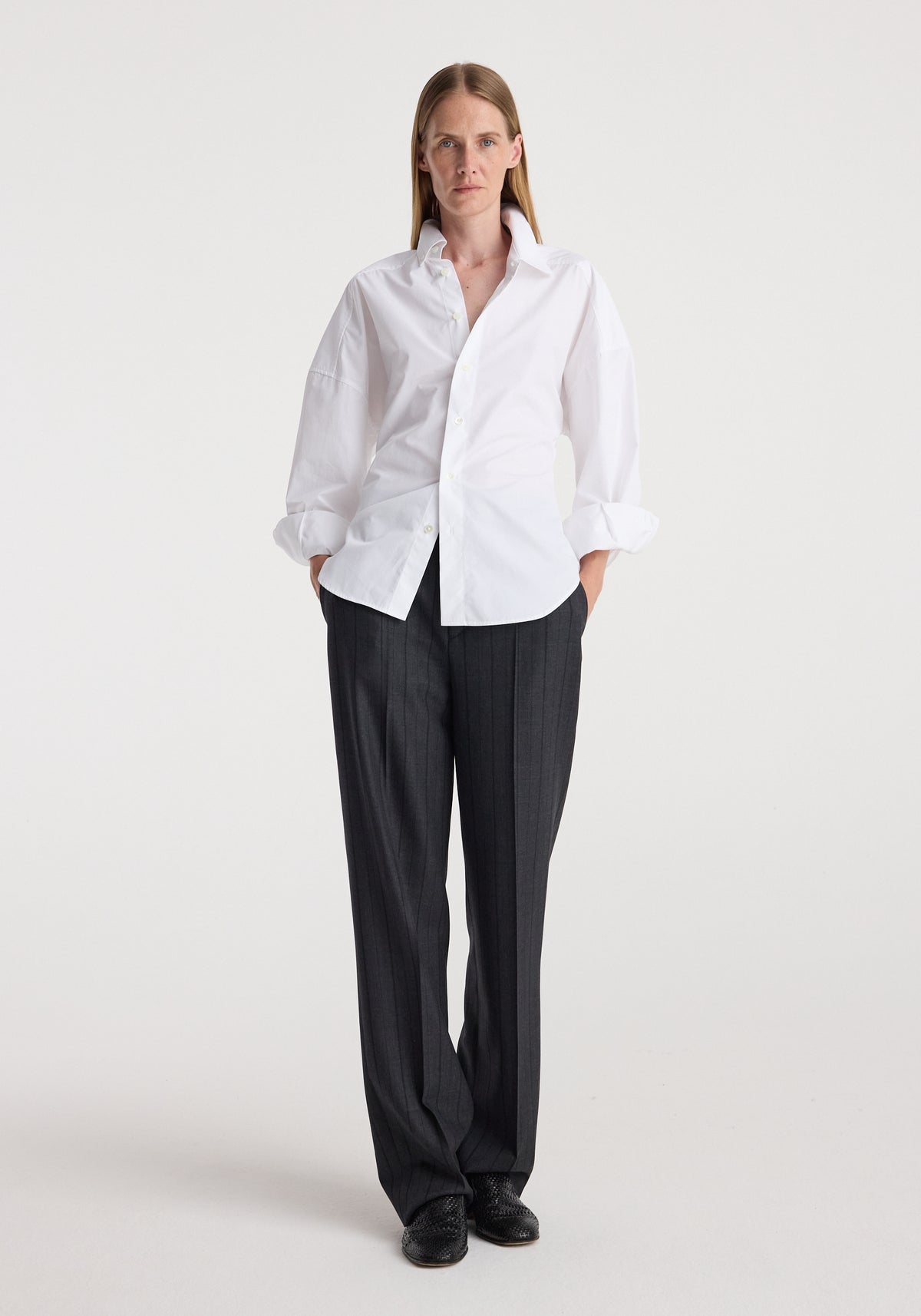 Woman wearing a white button-up shirt and Kee - Straight leg tailored trousers | charcoal stripe, standing against a plain background with hands in pockets and black shoes.