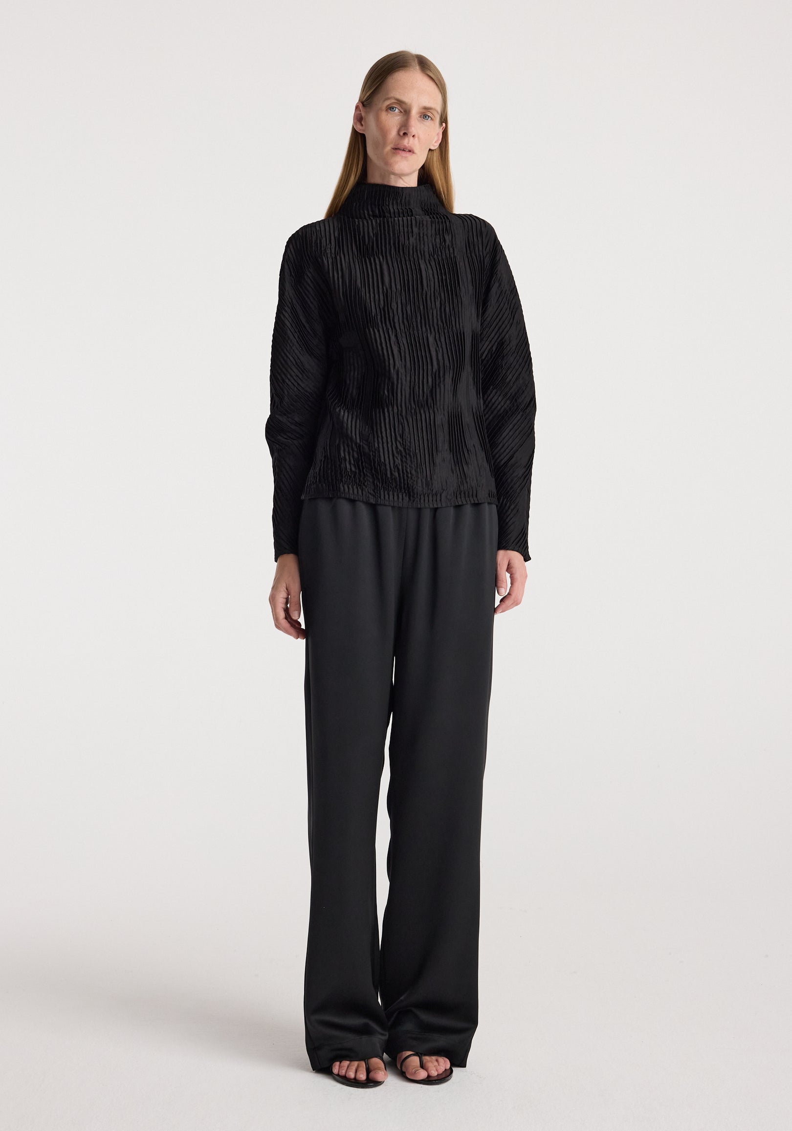 Woman wearing relaxed fit satin trousers | black paired with a textured black long-sleeve top, standing against a plain light background in a full-length view.
