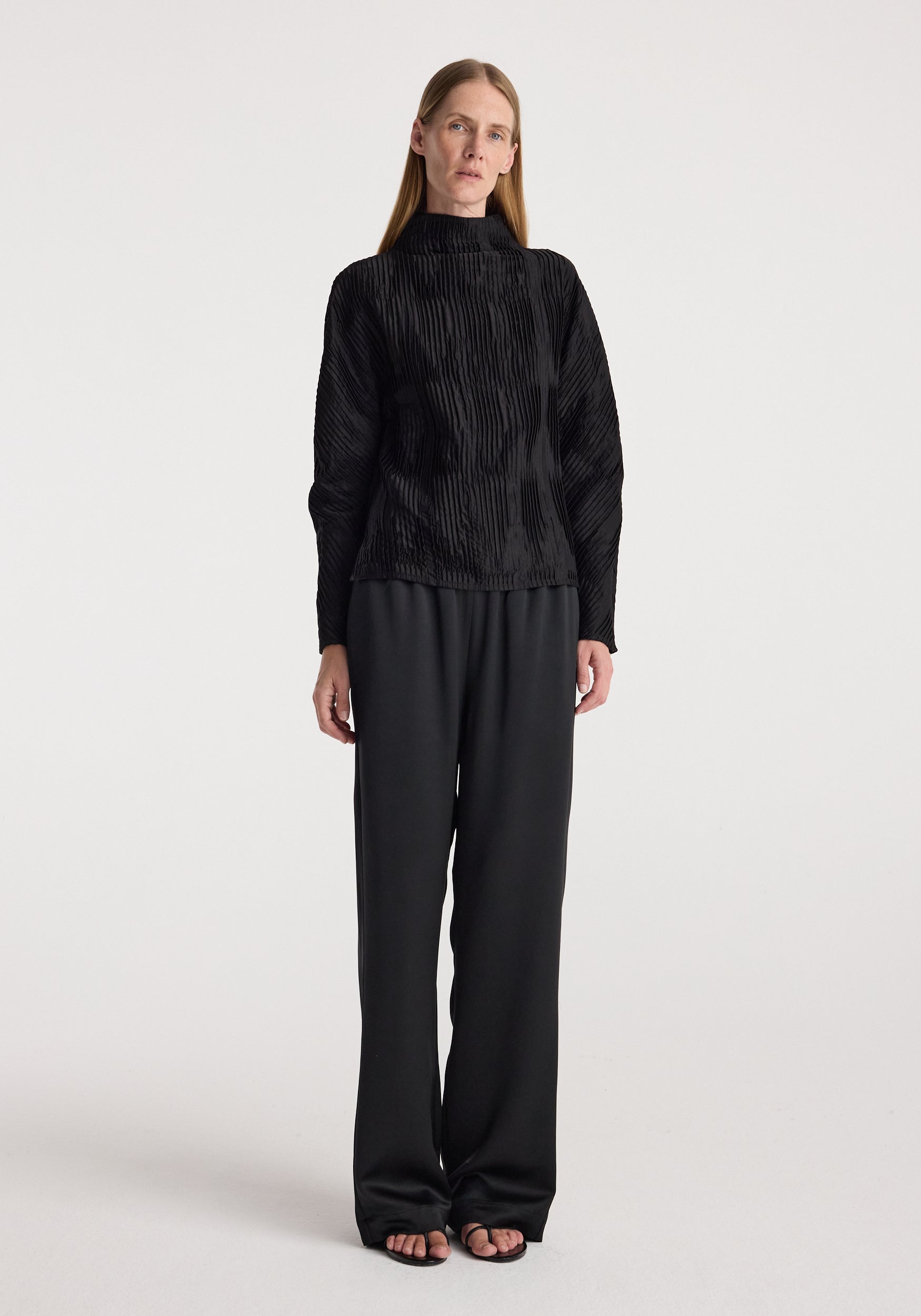 Woman wearing relaxed fit satin trousers | black paired with a textured black long-sleeve top, standing against a plain light background in a full-length view.