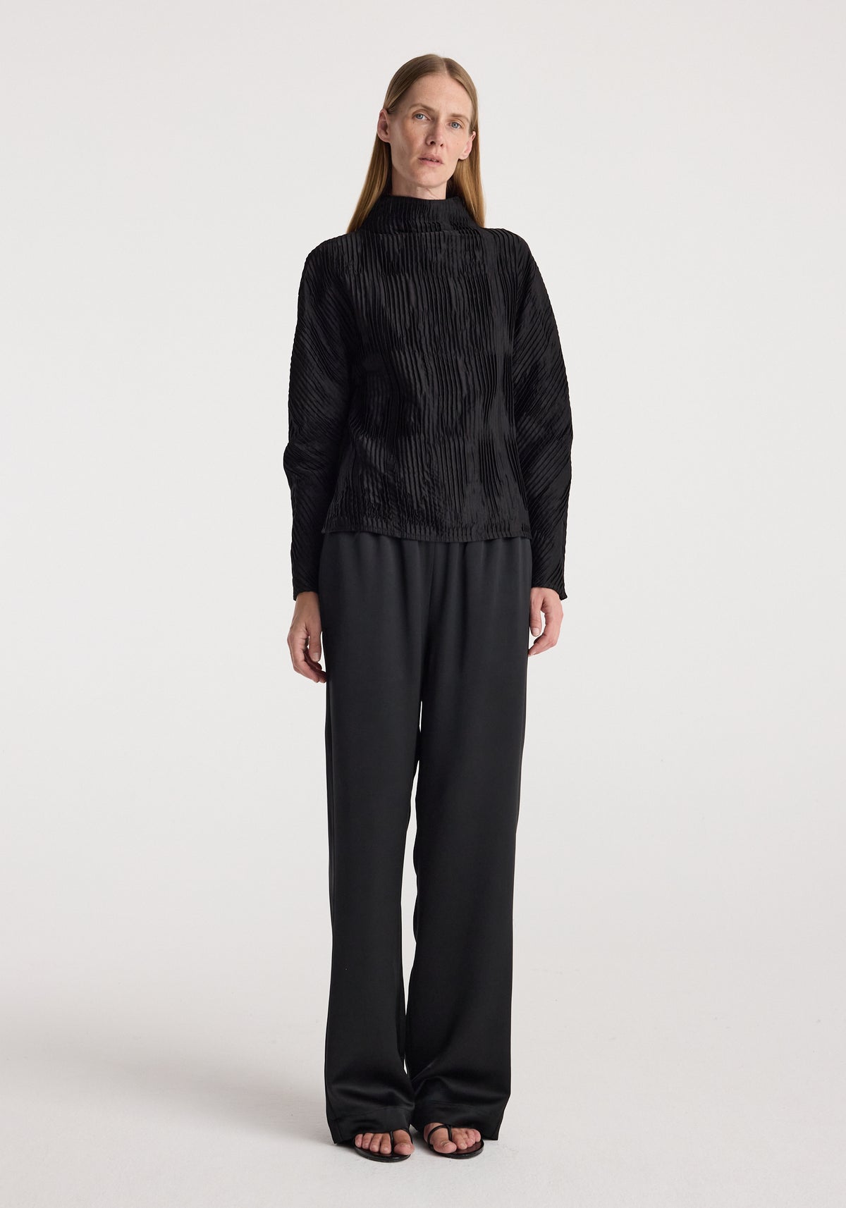 Woman wearing relaxed fit satin trousers | black paired with a textured black long-sleeve top, standing against a plain light background in a full-length view.