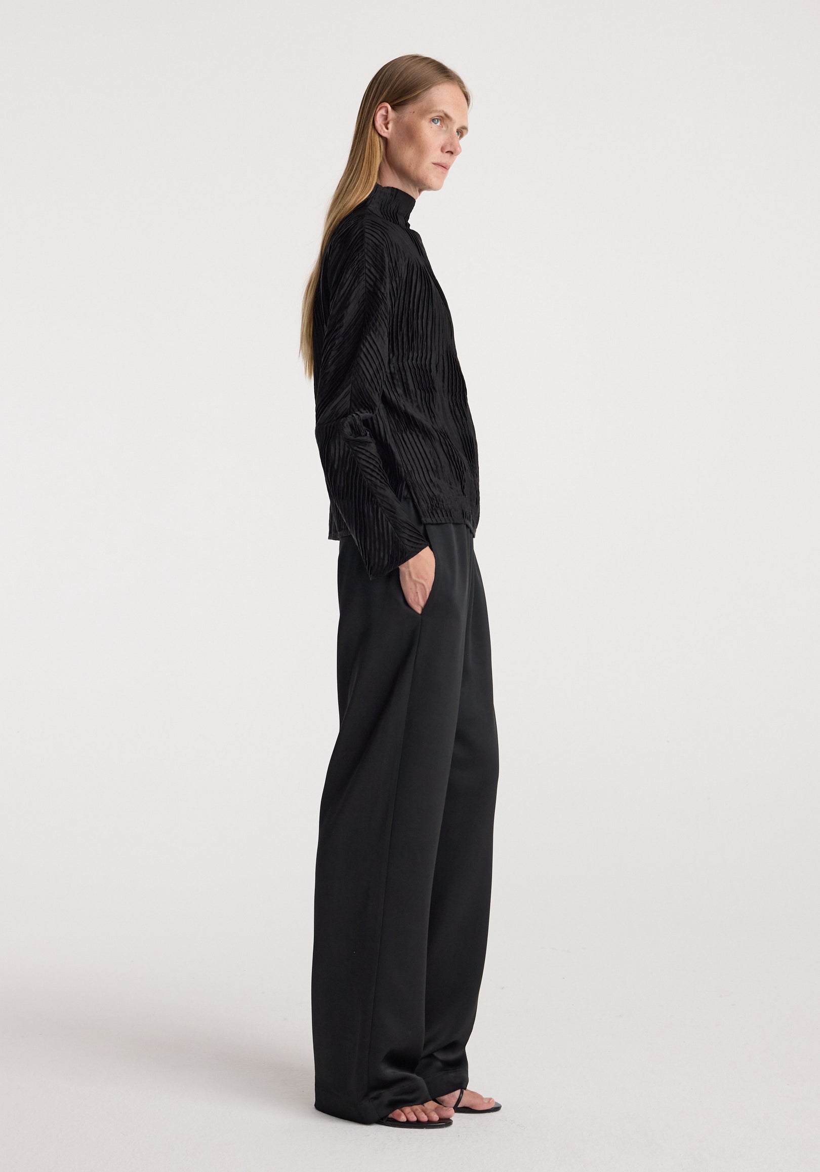 Side view of a woman wearing relaxed fit satin trousers | black paired with a long-sleeve textured black top, standing against a plain background with hands in pockets.