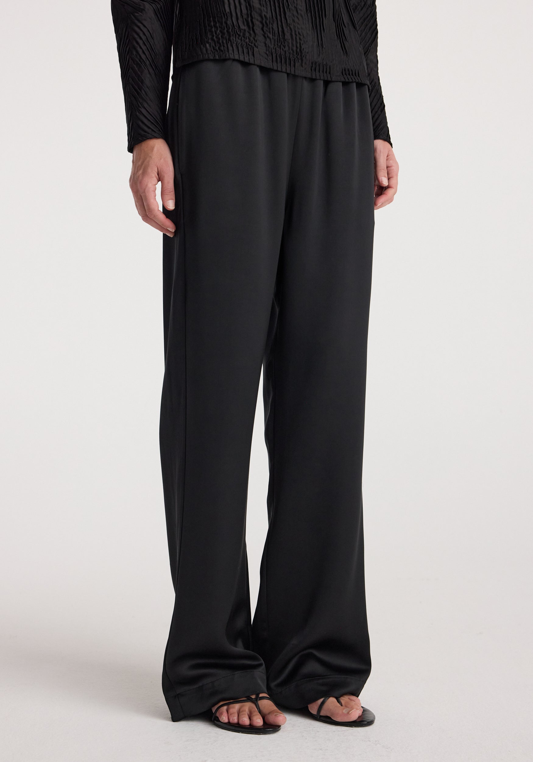 Front view of a person wearing relaxed fit satin trousers | black paired with a black top and black open-toe sandals against a plain light background.