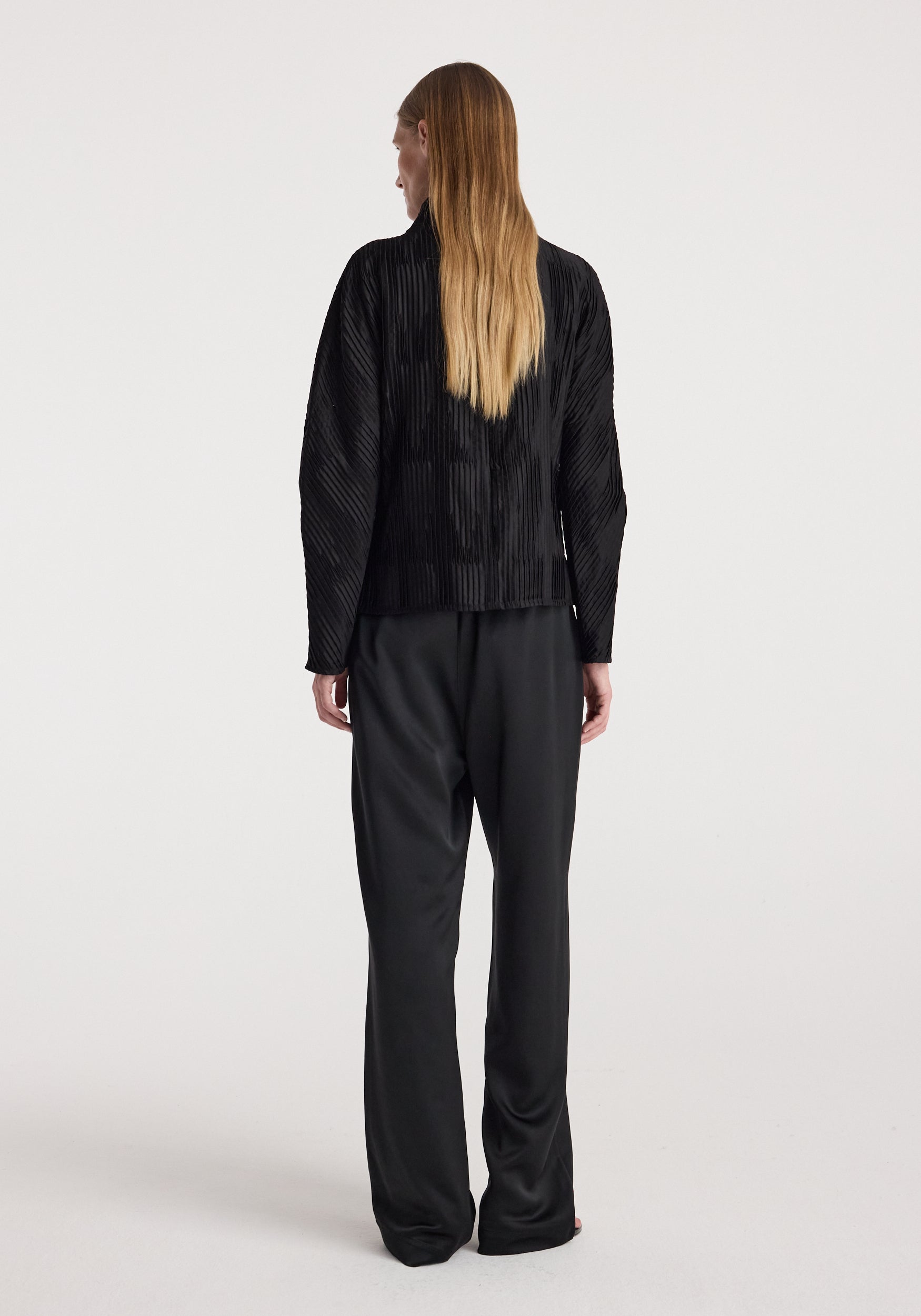 Woman seen from the back wearing relaxed fit satin trousers | black, paired with a long-sleeve black top, standing against a plain light background.