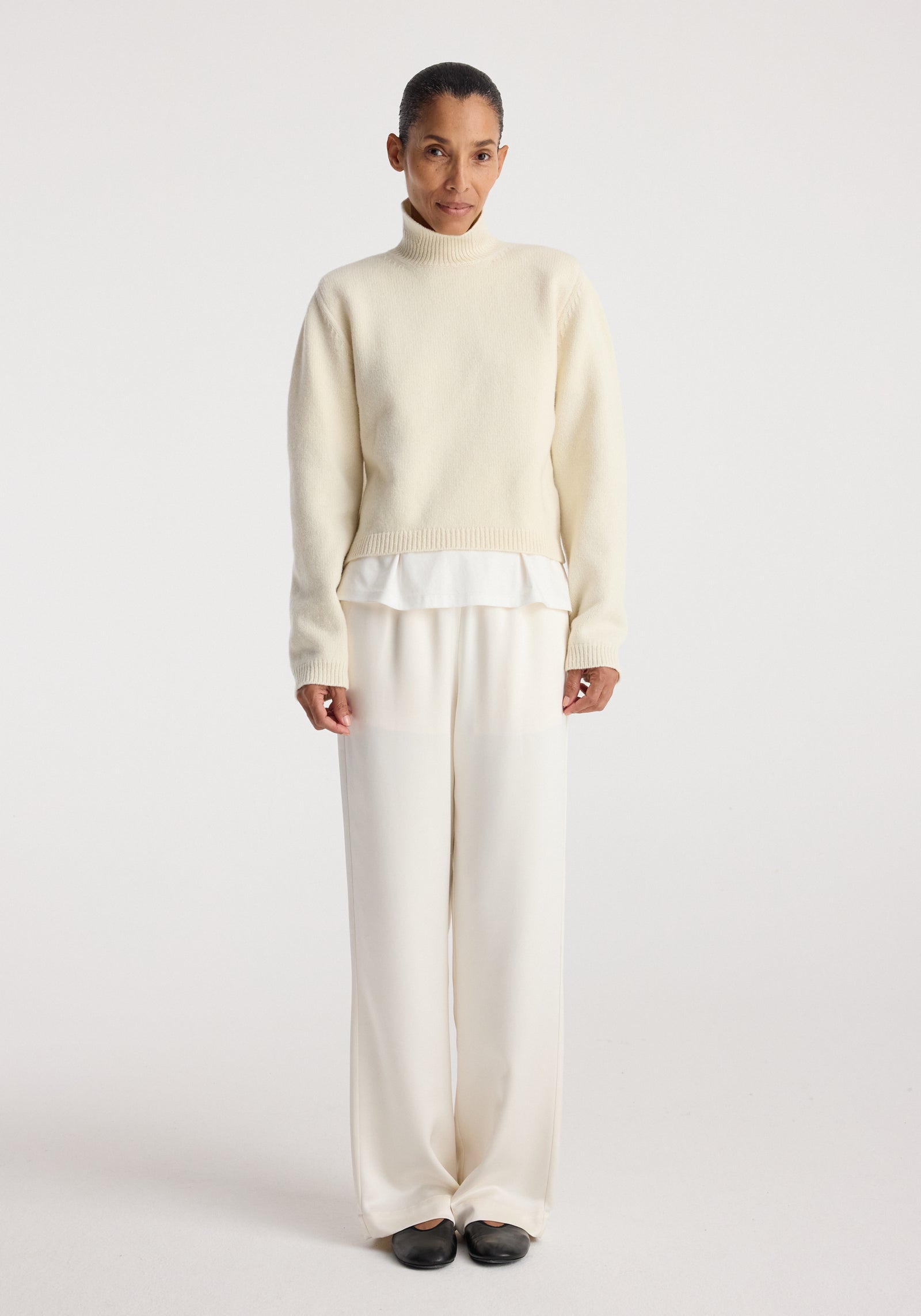 Woman wearing Relaxed fit satin trousers | cream paired with a cream sweater and black shoes, standing against a plain white background.