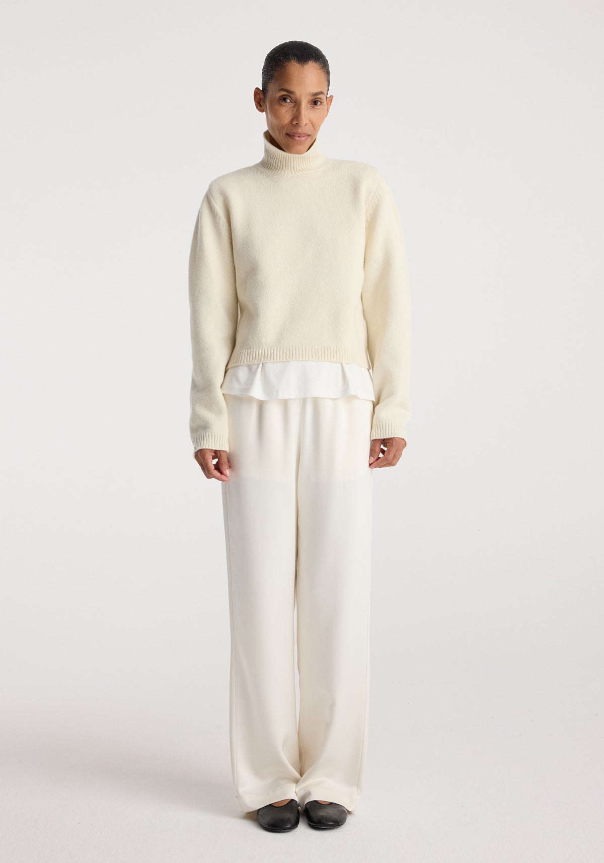 Woman wearing Relaxed fit satin trousers | cream paired with a cream sweater and black shoes, standing against a plain white background.