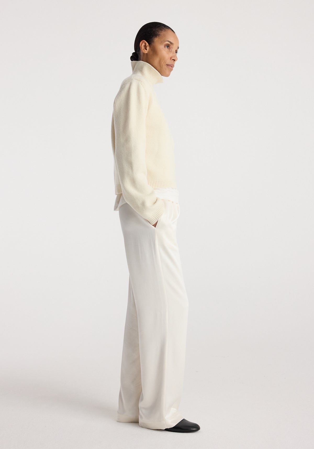 Woman wearing relaxed fit satin trousers | cream paired with a cream turtleneck sweater and black shoes, standing in profile against a plain background.