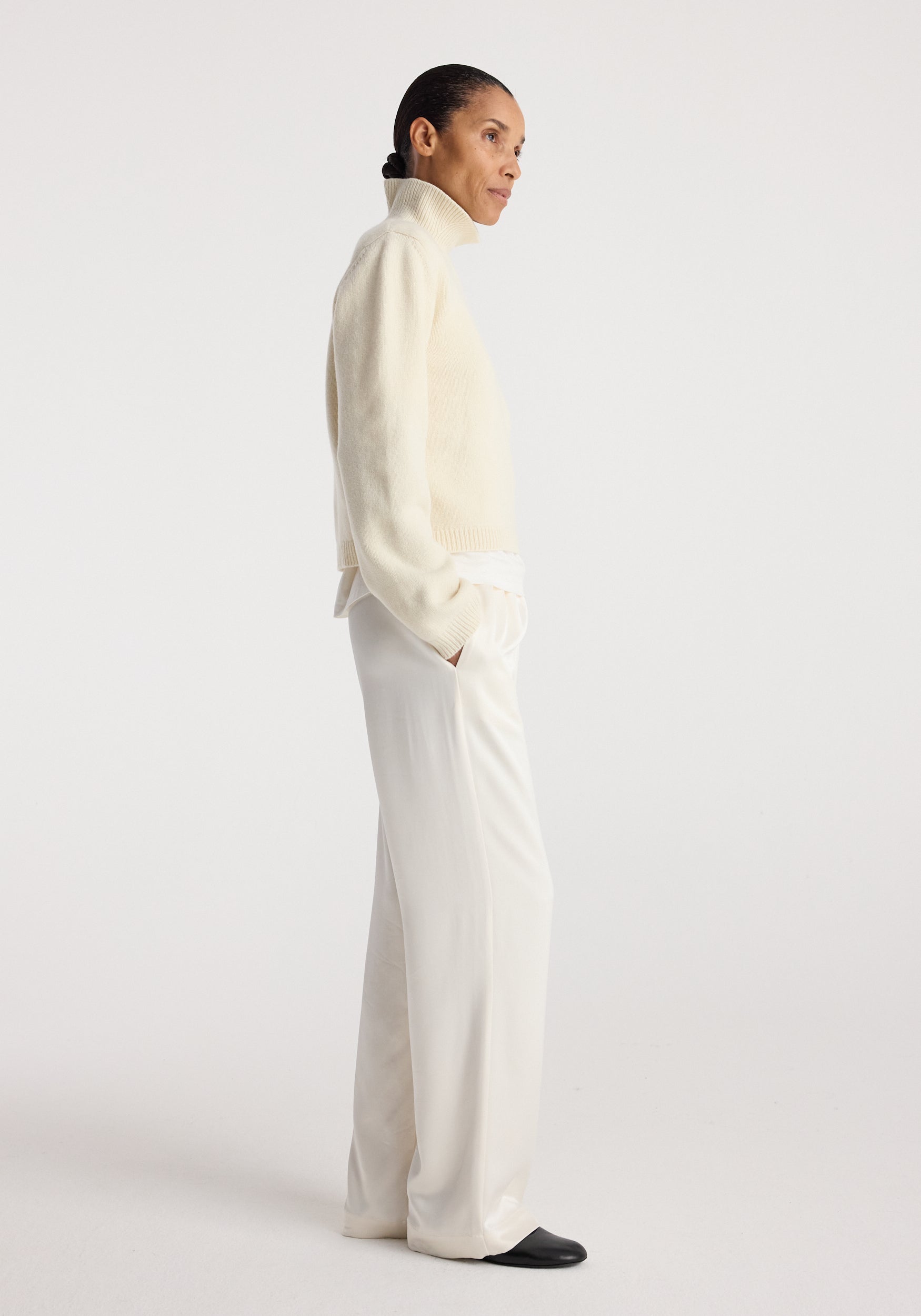 Woman wearing relaxed fit satin trousers | cream paired with a cream turtleneck sweater and black shoes, standing in profile against a plain background.