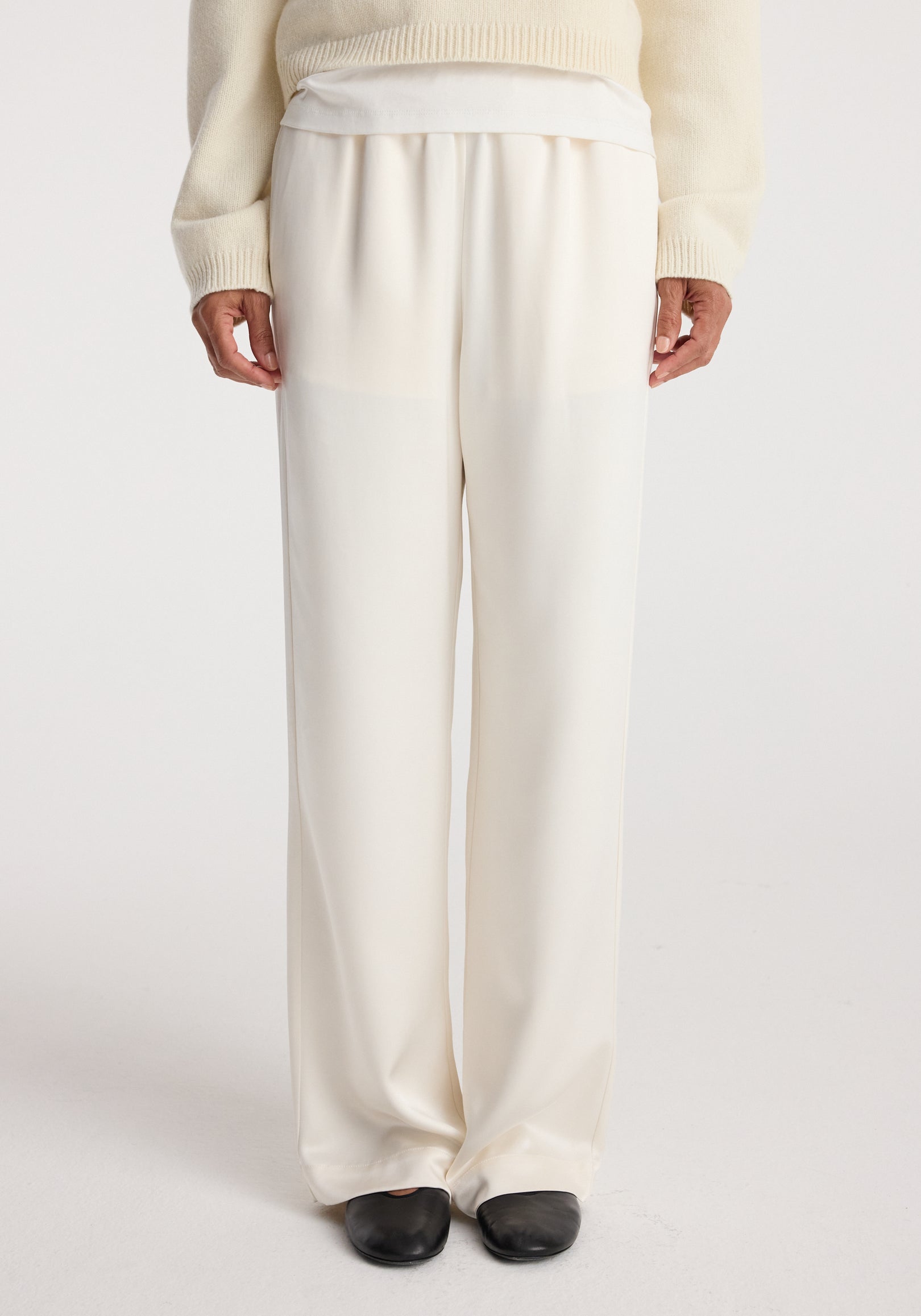 Front view of a person wearing relaxed fit satin trousers in cream with a loose silhouette, paired with a cream sweater and black shoes against a white background.