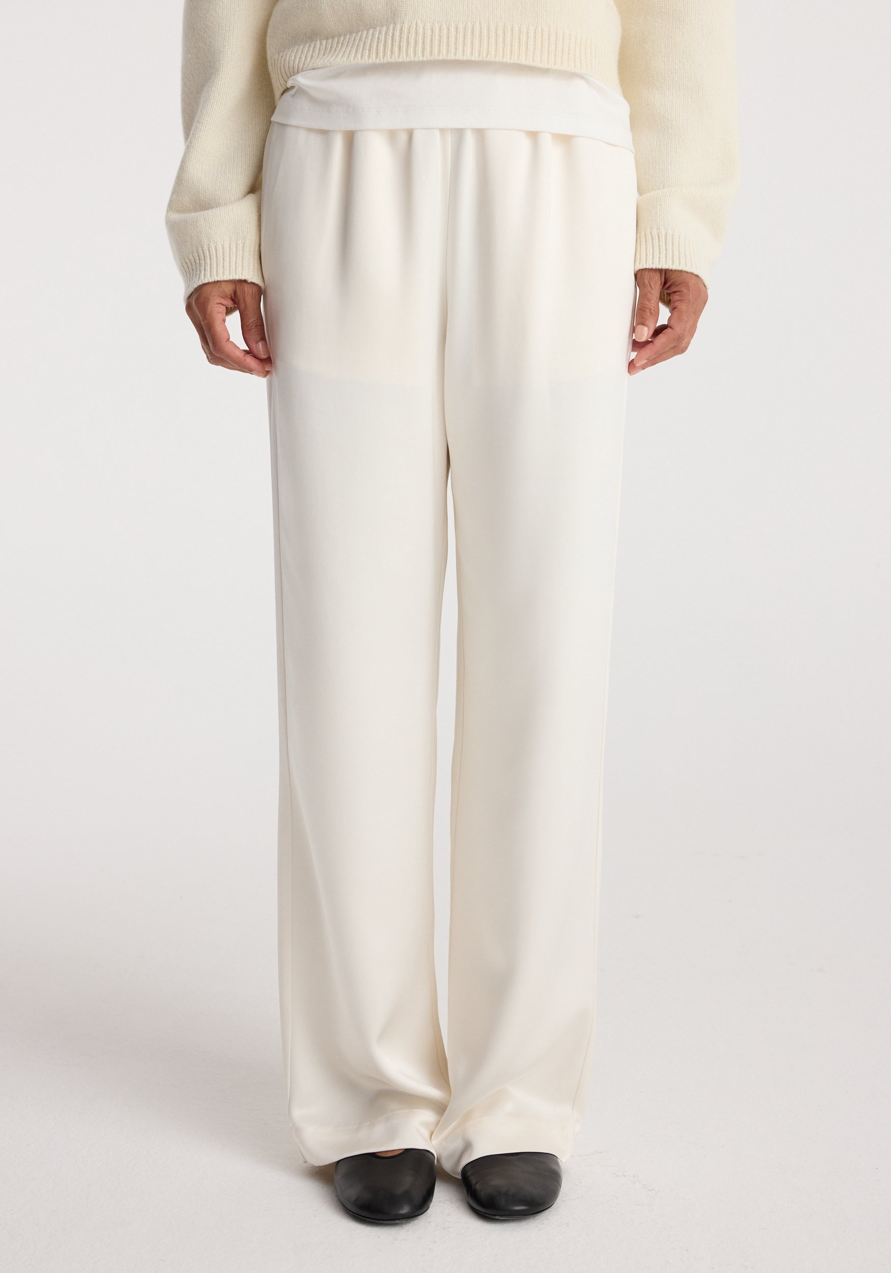 Front view of a person wearing relaxed fit satin trousers in cream with a loose silhouette, paired with a cream sweater and black shoes against a white background.