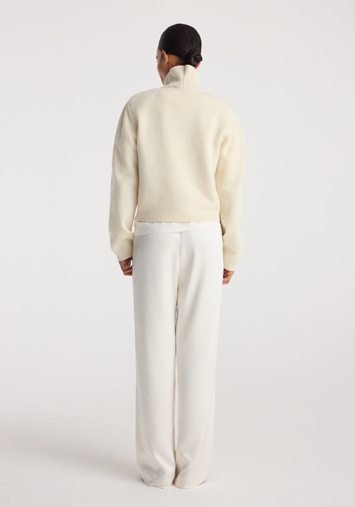 Woman wearing cream high-waisted Relaxed fit satin trousers | cream, paired with a cream turtleneck sweater, standing facing away against a plain background.