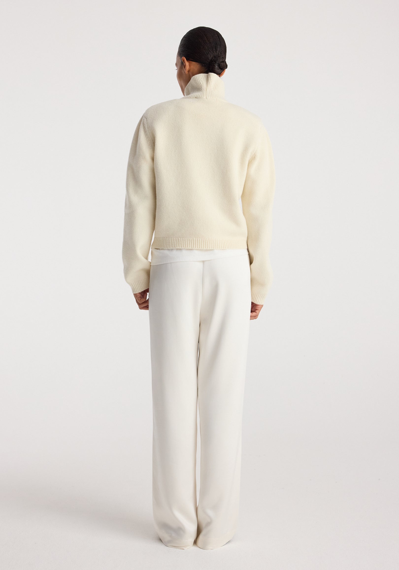 Woman wearing cream high-waisted Relaxed fit satin trousers | cream, paired with a cream turtleneck sweater, standing facing away against a plain background.