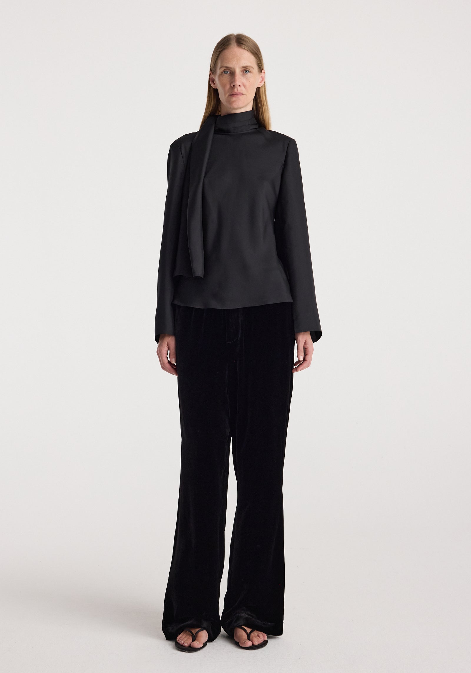 Woman wearing black long-sleeve top paired with Velvet relaxed trousers | black and black open-toe sandals, standing against a plain white background.