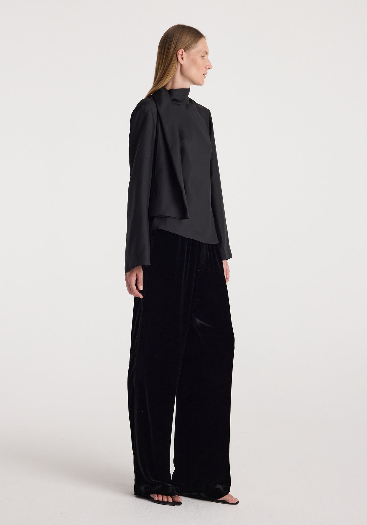 Woman wearing black velvet relaxed trousers | black paired with a long-sleeve black top, standing against a plain light background in a side profile pose.