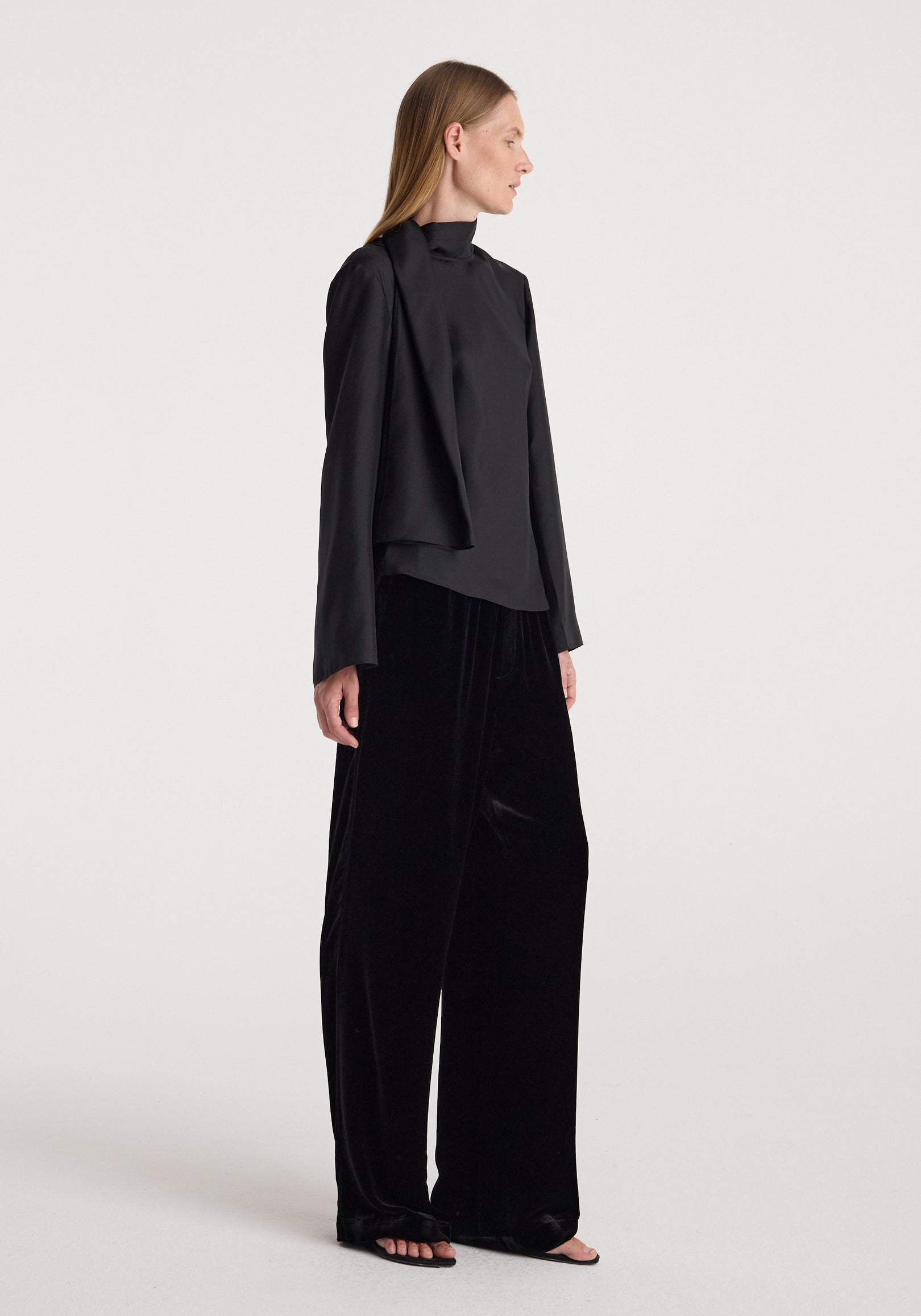 Woman wearing black velvet relaxed trousers | black paired with a long-sleeve black top, standing against a plain light background in a side profile pose.