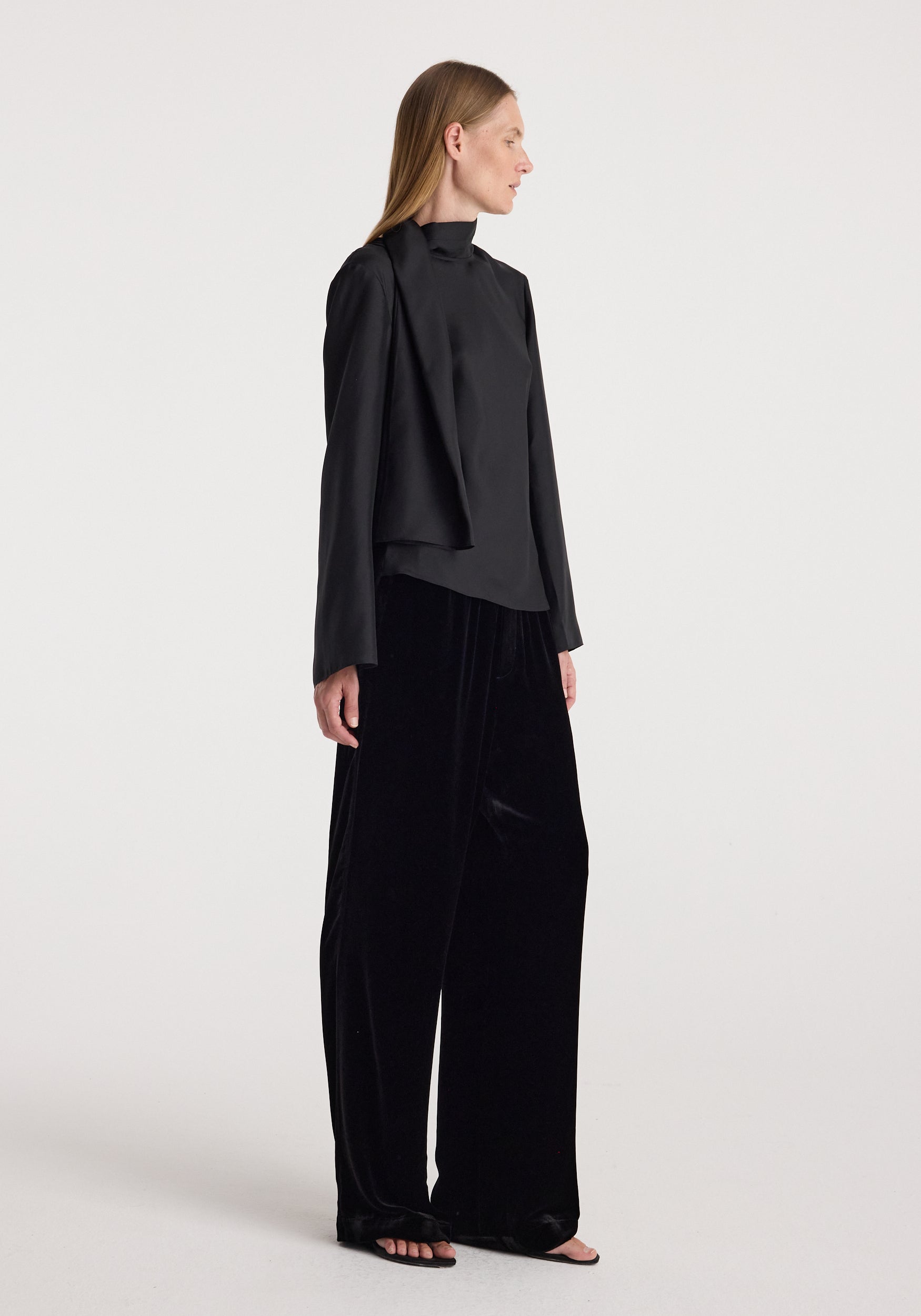 Woman wearing black velvet relaxed trousers | black paired with a long-sleeve black top, standing against a plain light background in a side profile pose.