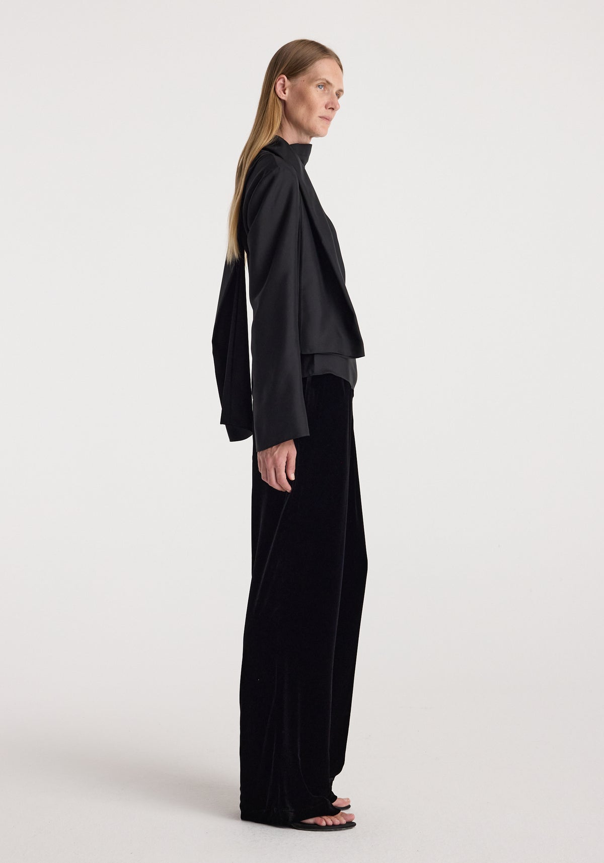 Woman wearing black velvet relaxed trousers | black paired with a loose black long-sleeve top, standing in profile against a plain light background.
